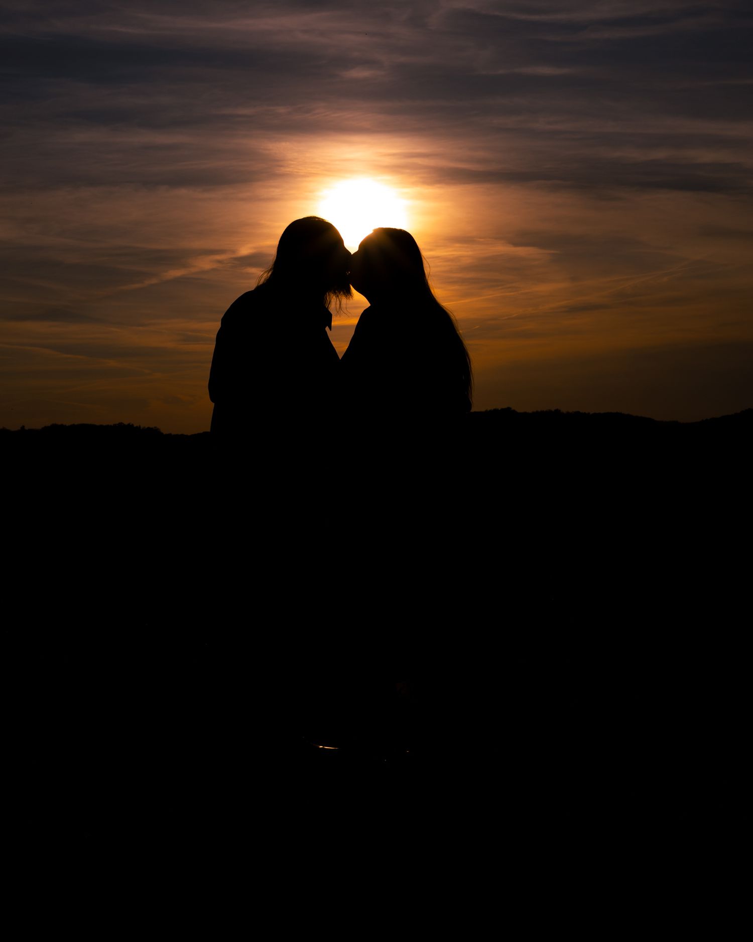 Romantic silhouette of couple kissing at golden sunset, forming heart shape with glowing sun behind them.