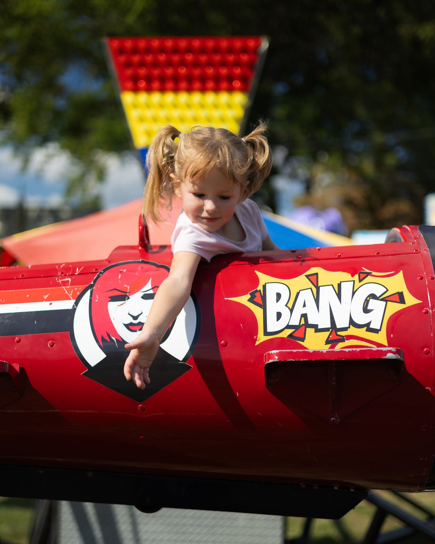 Young girl with pigtails playing in a red BANG carnival ride at an outdoor fair.