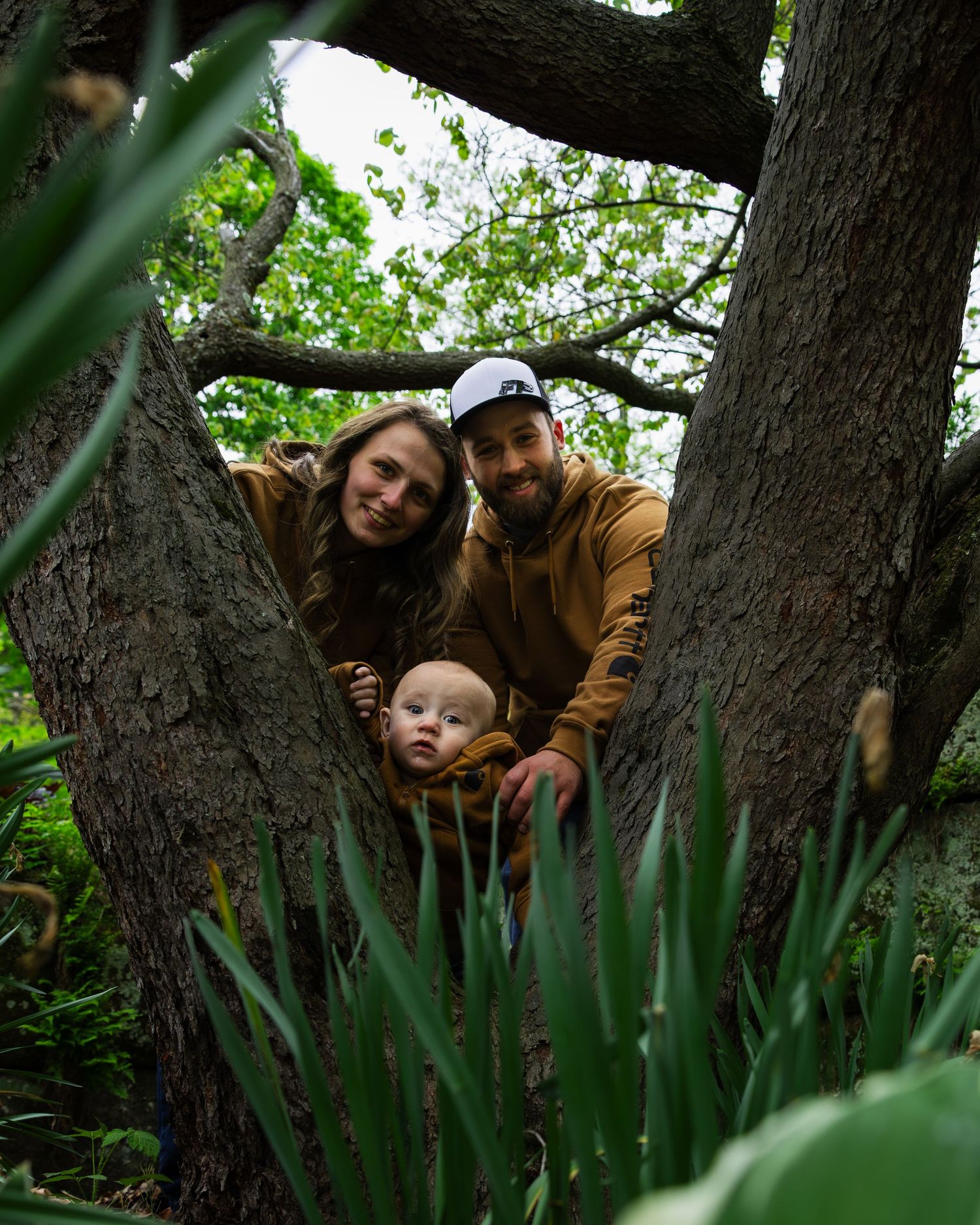 Happy couple with their baby peek through tree branches in a lush green garden setting.