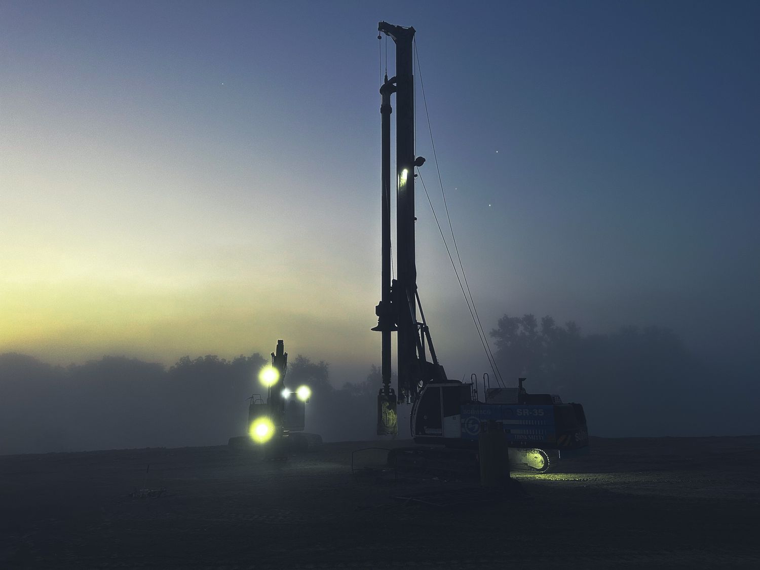 Large drilling rig with bright lights operating at dusk in misty conditions on an open construction site.