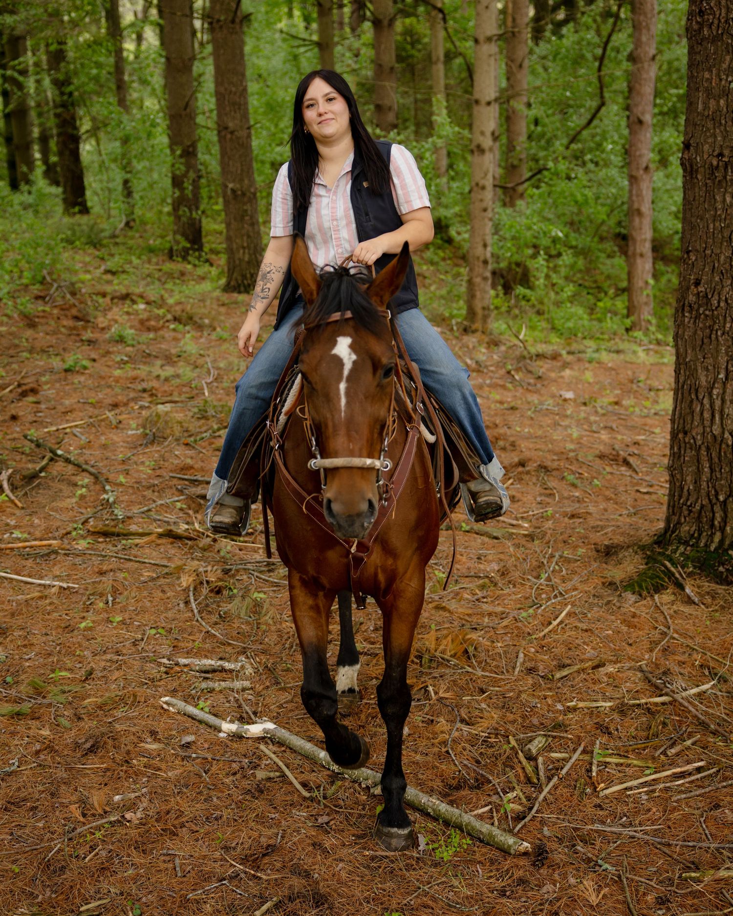 Woman with long dark hair rides a brown horse through a forest trail surrounded by pine trees and fallen branches.