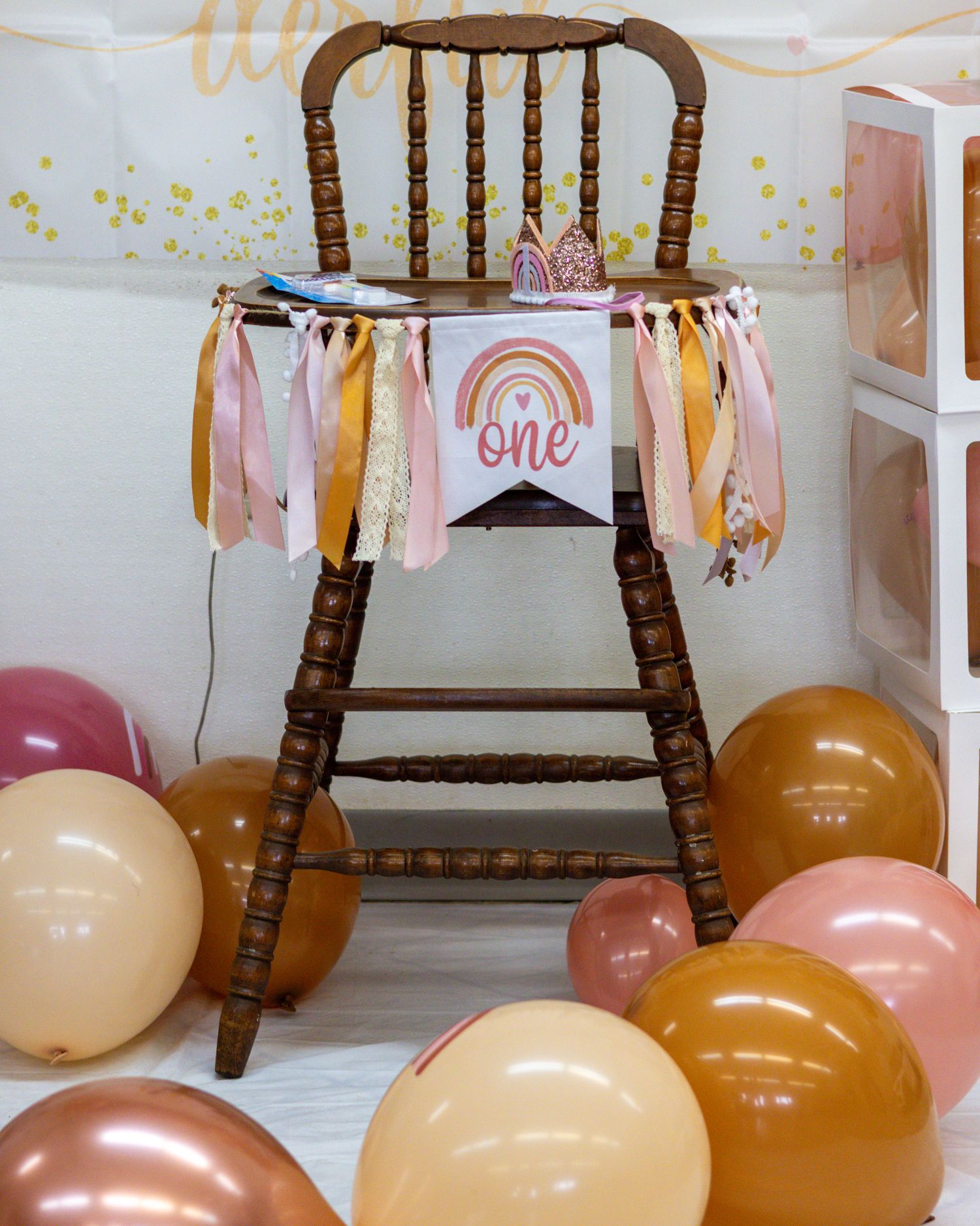 First birthday high chair decorated with rainbow one banner and colorful balloons on floor.