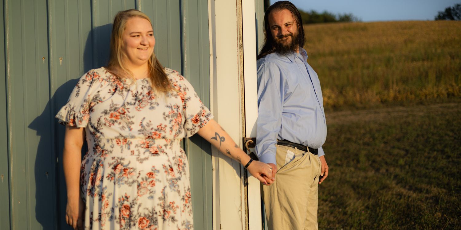 Couple posing outdoors at golden hour near a barn, woman in floral dress and man in blue shirt holding hands.