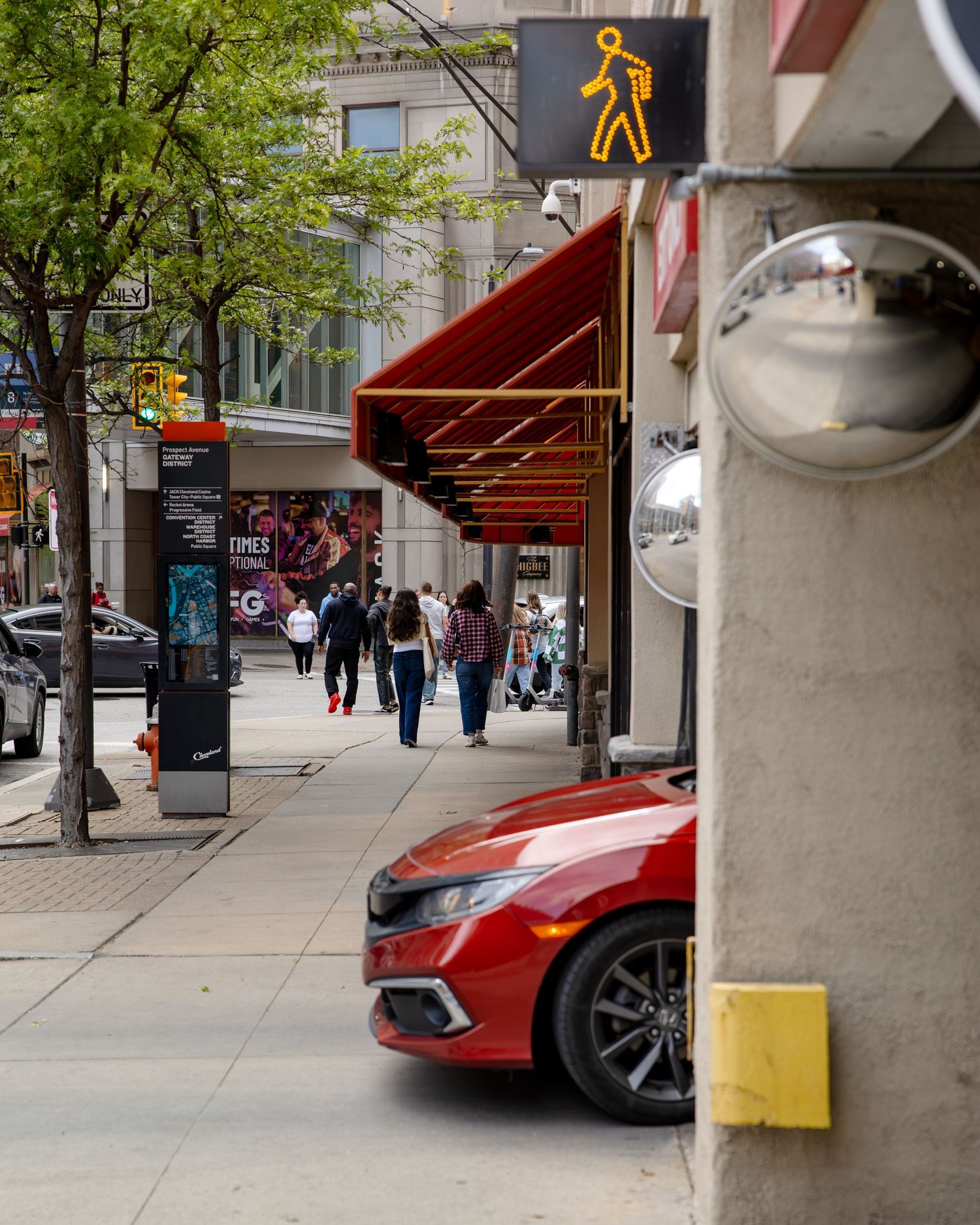 Urban sidewalk scene with pedestrian crossing sign, red Honda Civic, and people walking past red awning storefronts.