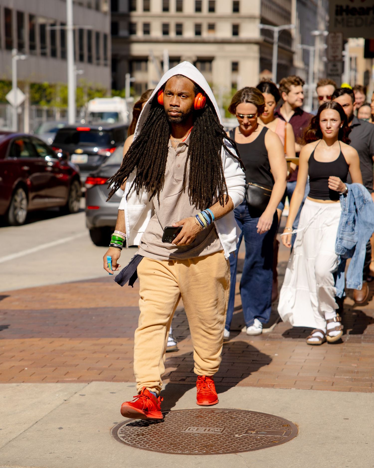 Man with long dreadlocks wearing orange headphones, white hoodie, tan pants, and red sneakers walking on a city sidewalk.