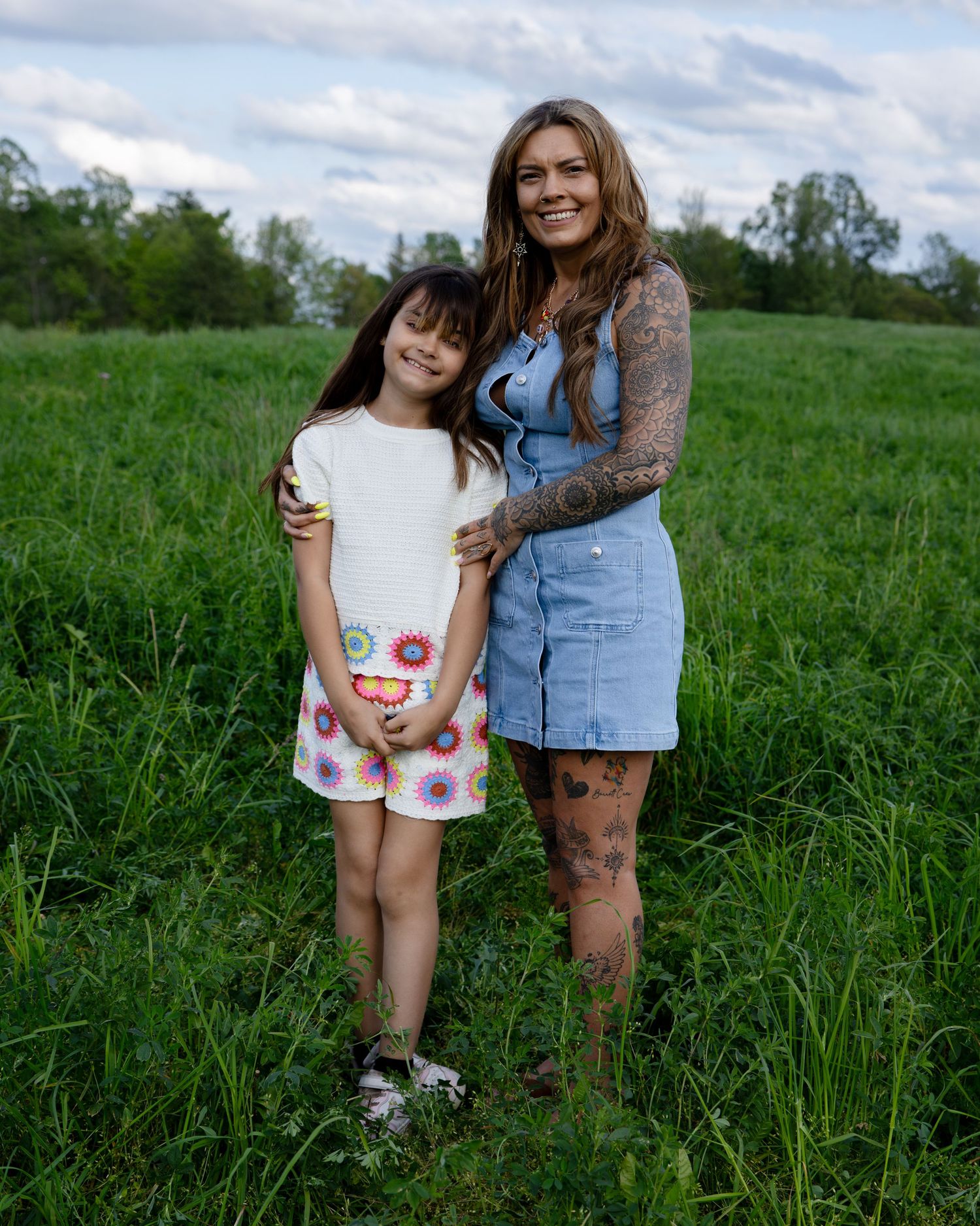 Woman and young girl posing together in a green field, smiling, with trees and cloudy sky in the background.