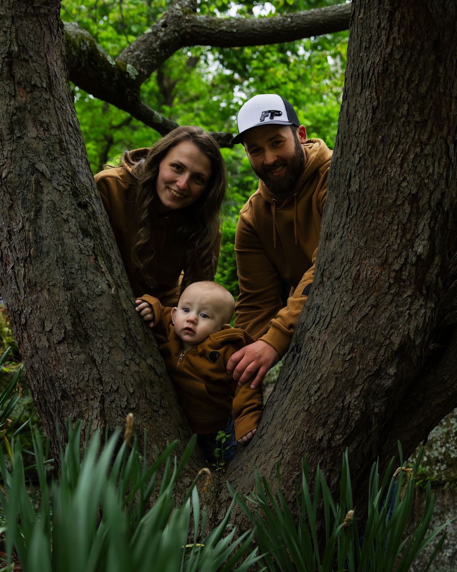 Happy young family with baby poses between large tree trunks in lush green botanical garden.