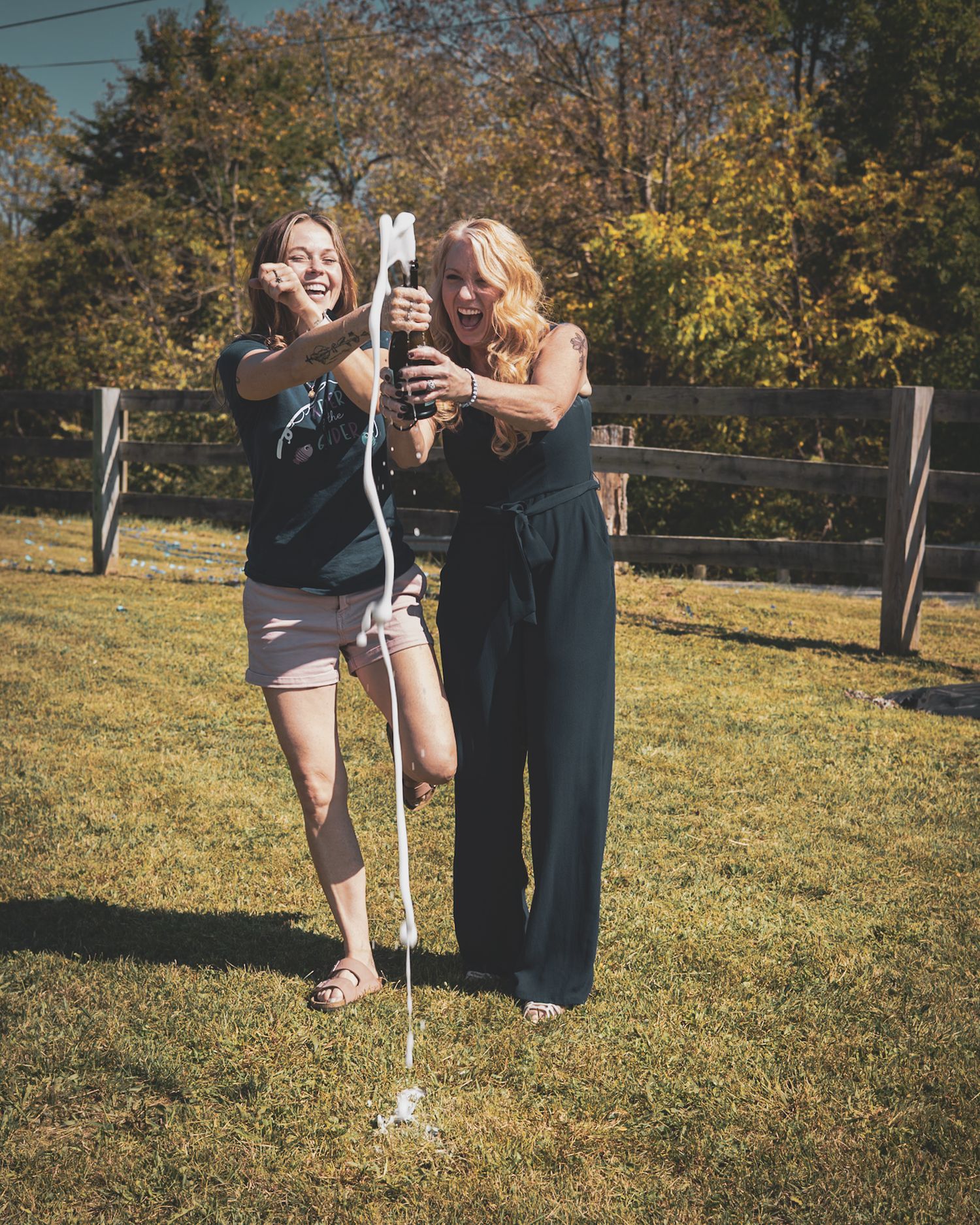 Two women laughing while using a confetti popper at an outdoor celebration on a sunny day.