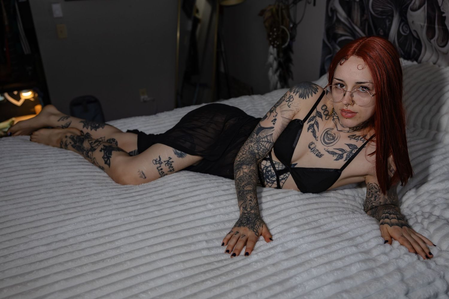 Tattooed redhead woman in black mesh dress lying on white bed looking at camera.