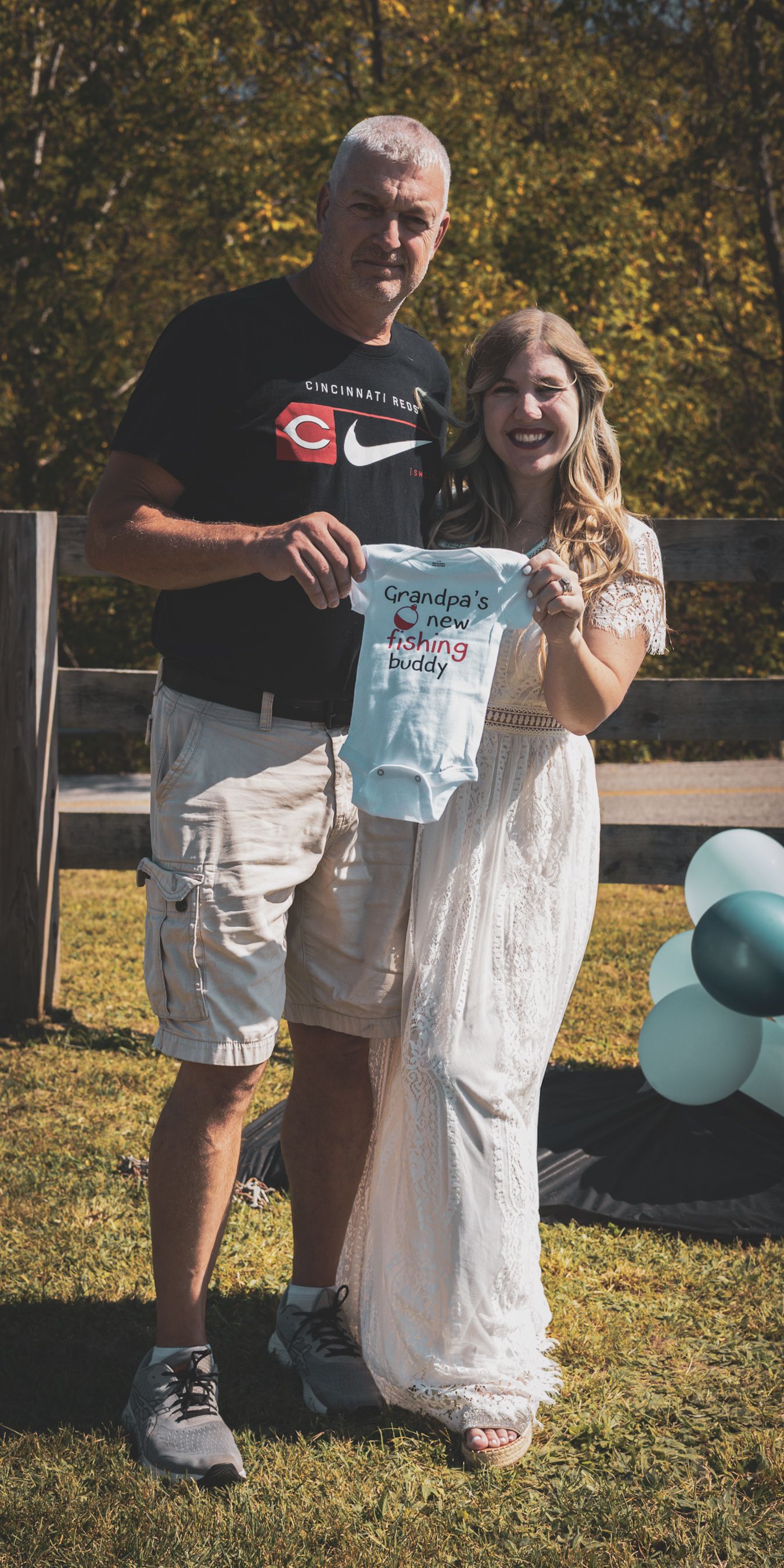 Proud grandfather and pregnant woman hold a blue Grandpas New Fishing Buddy onesie at a gender reveal party.
