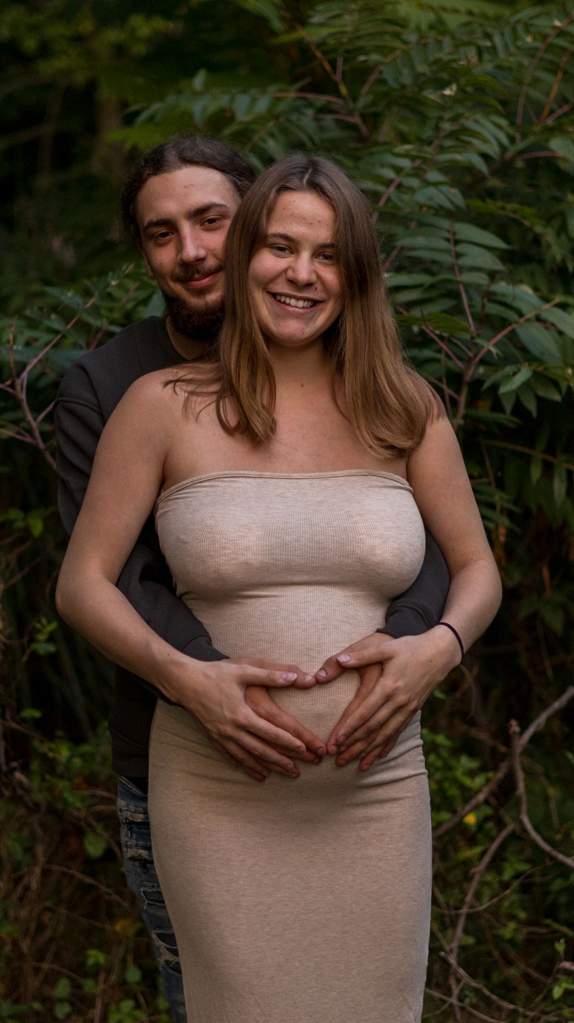 Smiling pregnant woman in beige tube dress poses with partner who embraces her from behind outdoors.