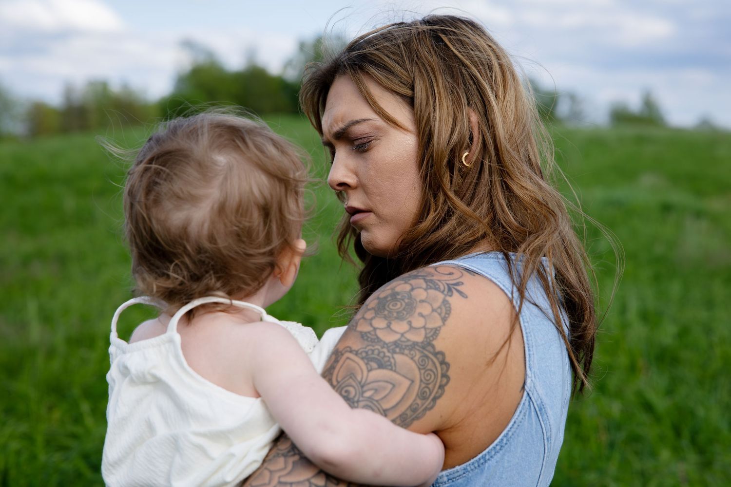 Tattooed young mother holding baby in white dress in green field.