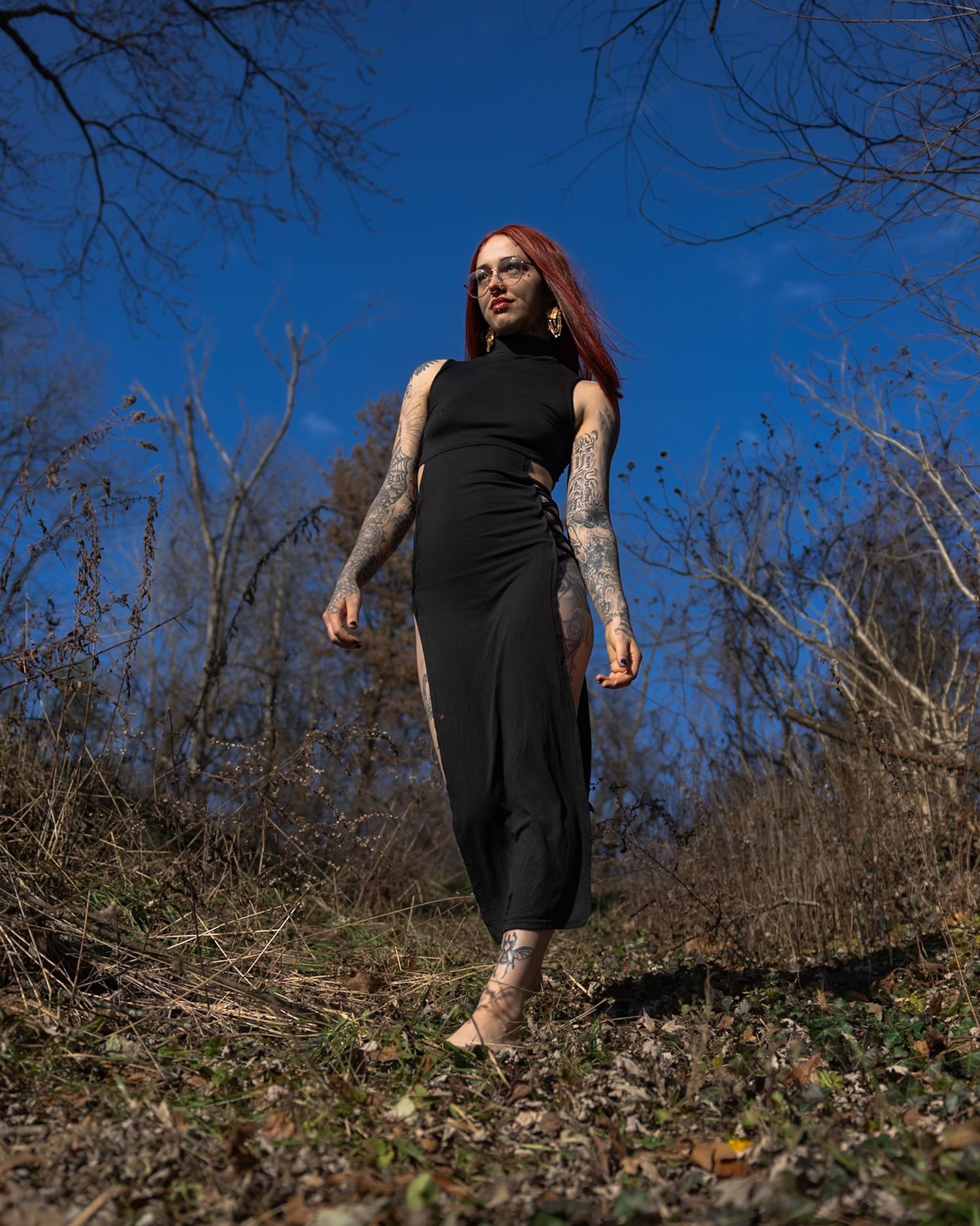 Tattooed redhead woman in black maxi dress walking through bare winter trees under blue sky.