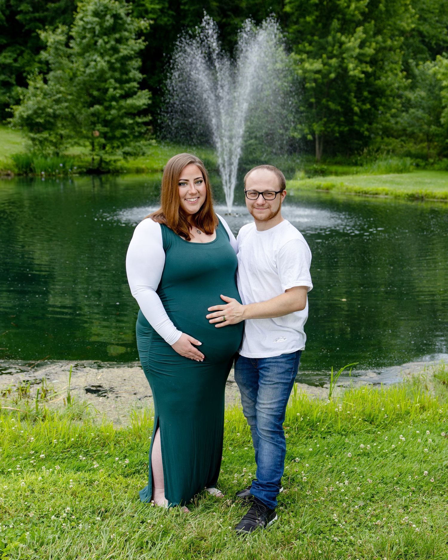 Pregnant couple posing by a serene pond with a fountain, surrounded by lush greenery, woman in green dress.
