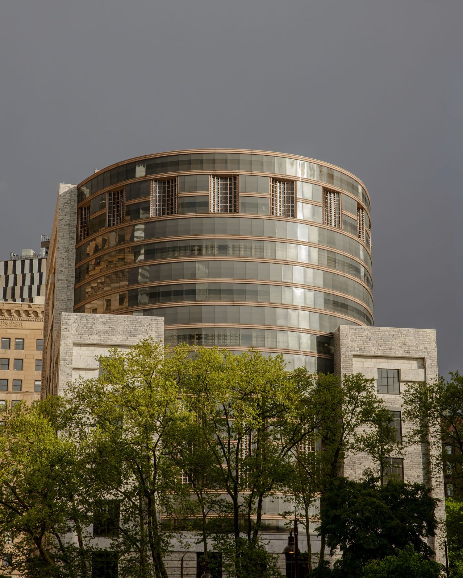 Curved glass skyscraper reflecting city buildings under dramatic stormy sky, surrounded by green trees in urban setting.