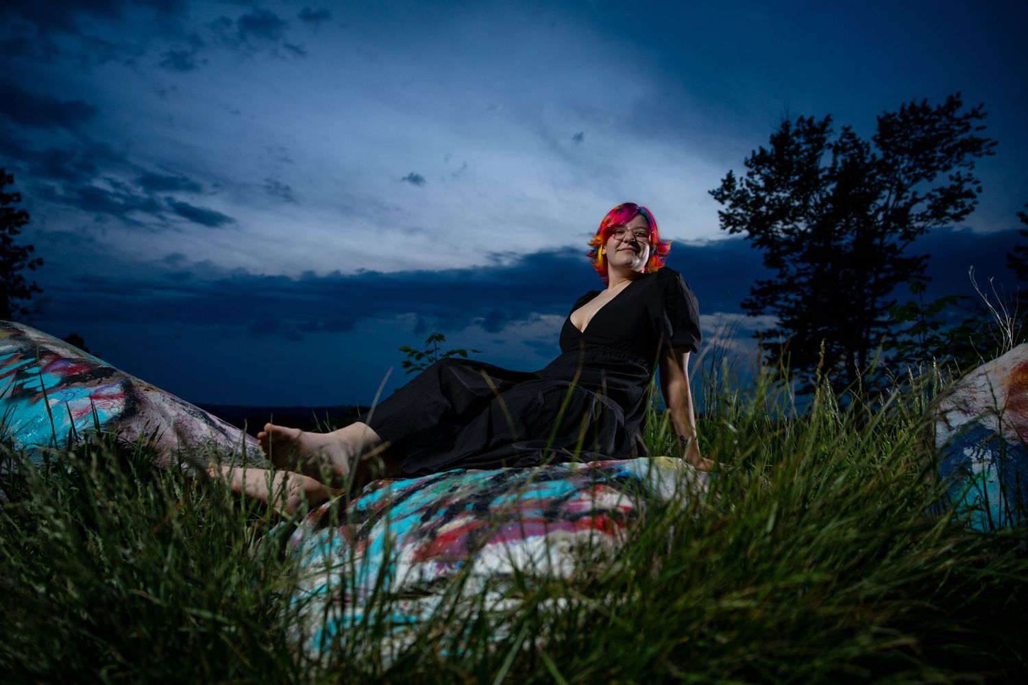 Woman with pink hair relaxing outdoors at dusk, surrounded by green grass and colorful graffiti under dramatic blue sky.