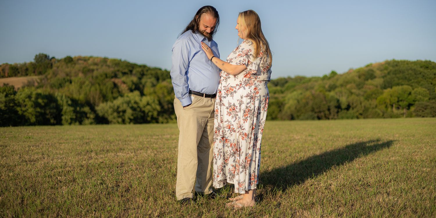 Pregnant couple sharing a tender moment in a sunny green field with trees in the background.