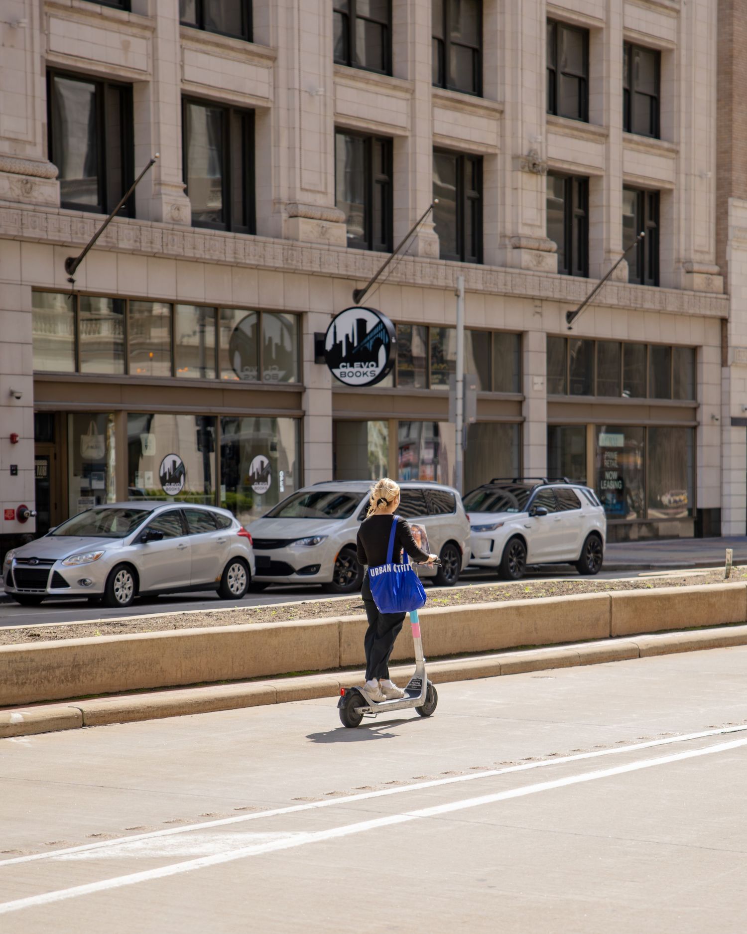 Woman riding electric scooter on urban street with parked cars and large city building in background.
