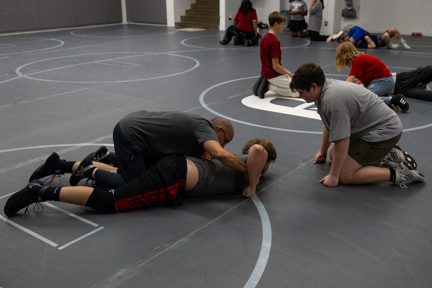 Young wrestlers practicing takedown techniques on a gray mat in an indoor gym.