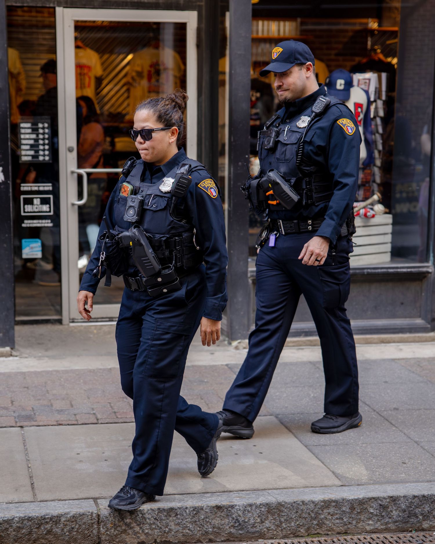 Two police officers in navy uniforms patrolling a city sidewalk past storefronts.