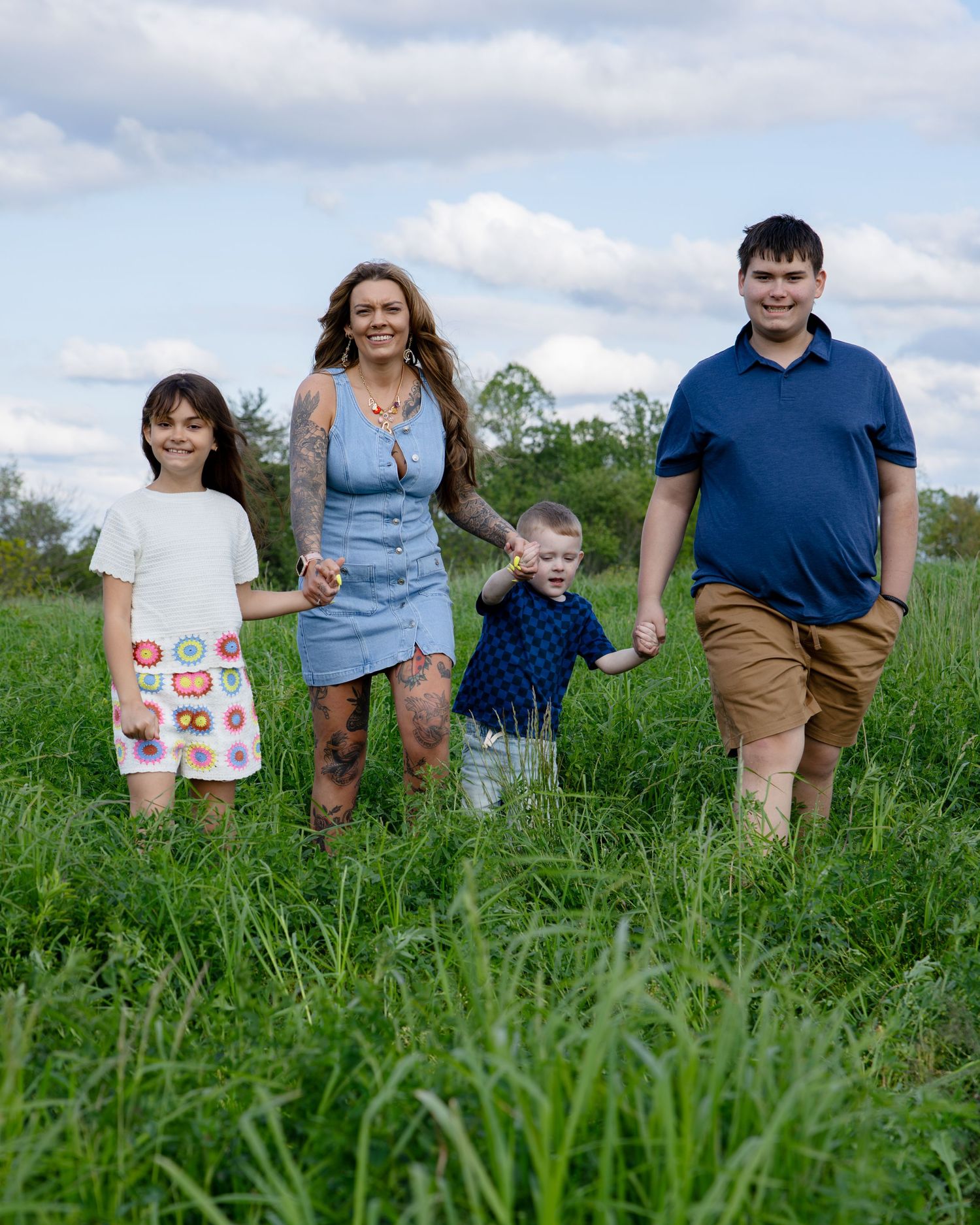 A tattooed mom walks hand-in-hand with her three children through a lush green field under a cloudy sky.
