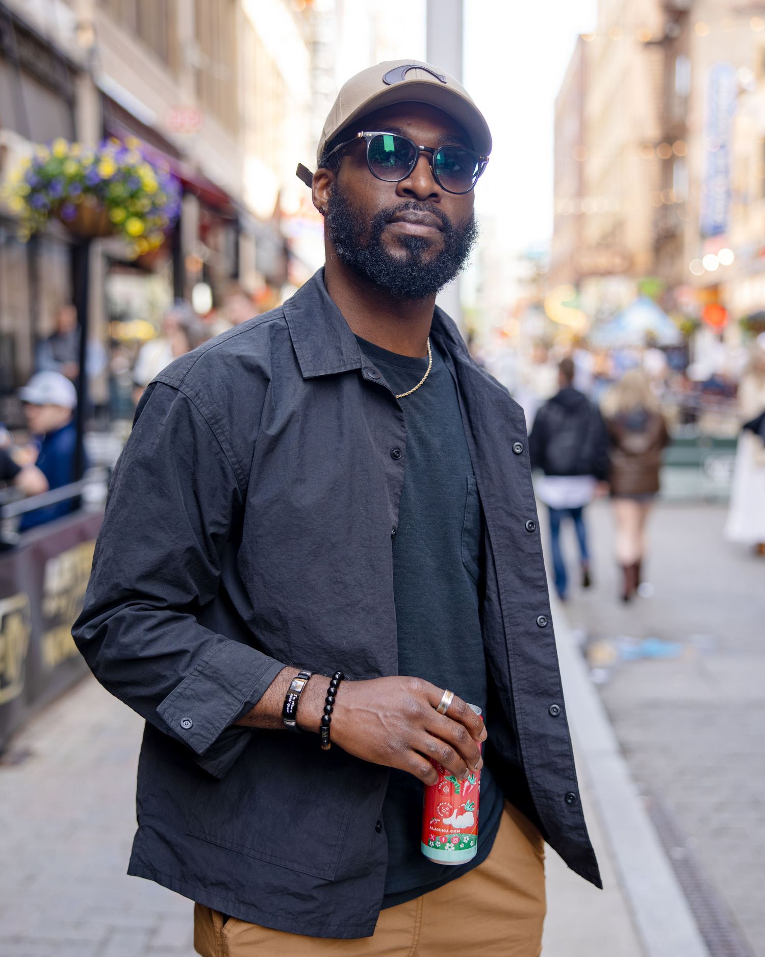 A stylish man in a dark jacket, cap, and sunglasses holds a can while standing on a busy urban street.