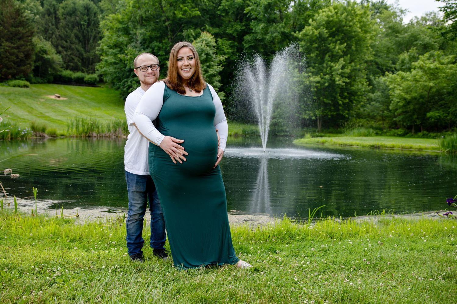Pregnant woman in green gown with partner by a pond and fountain in a lush park maternity photo.