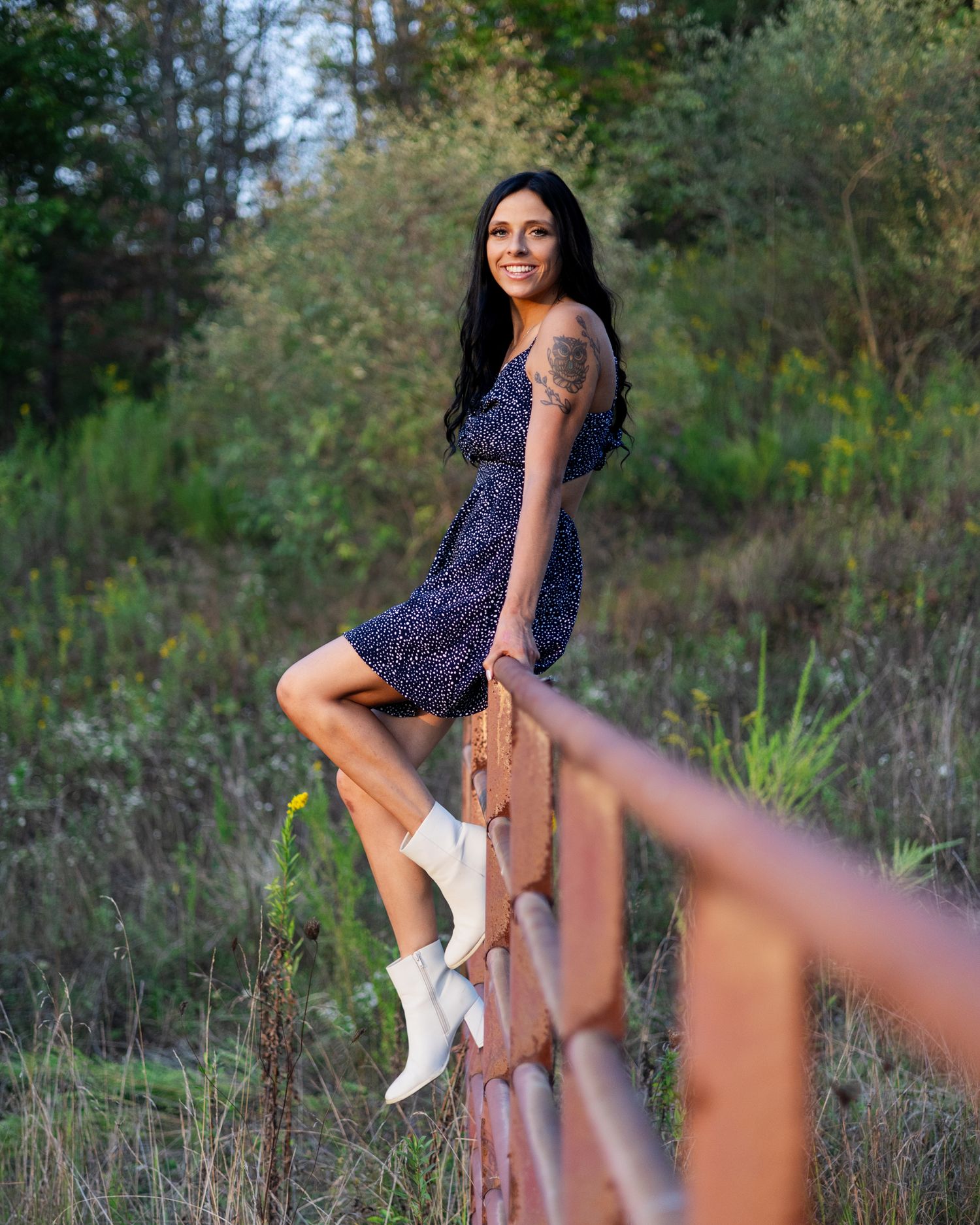 Woman in floral mini dress and white boots posing on rustic fence in nature.