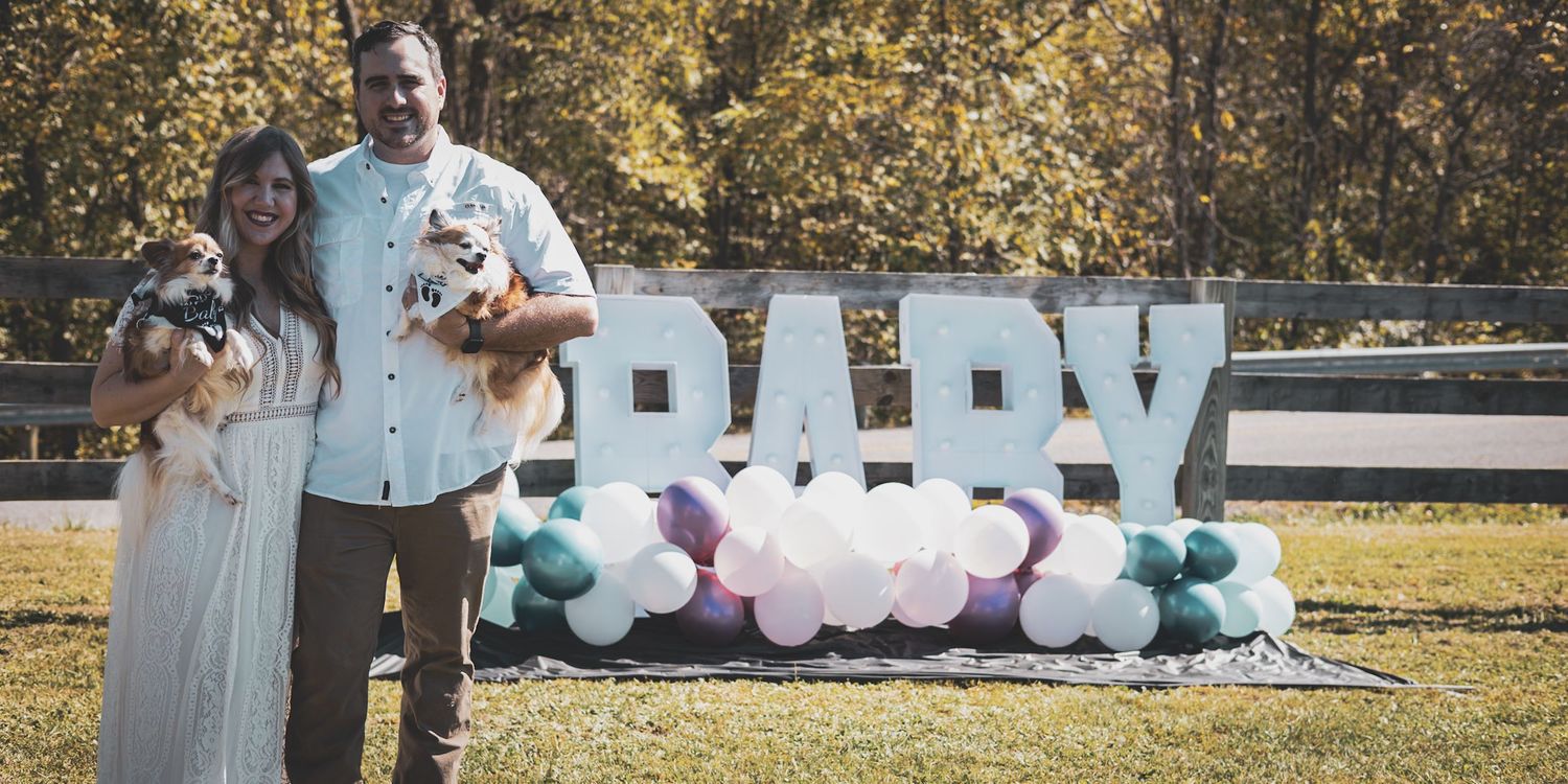 Expecting couple holding dogs in front of BABY balloon display at gender reveal party.