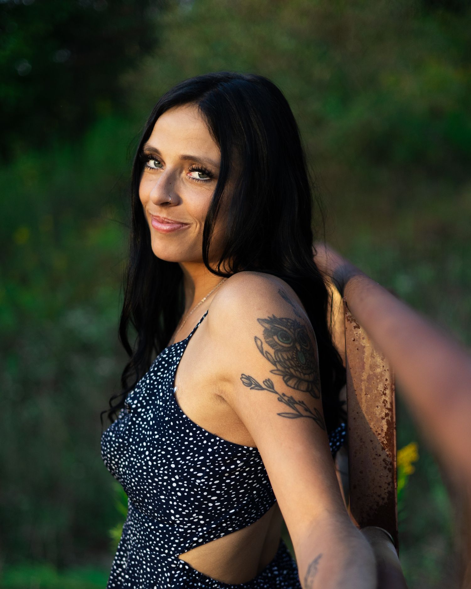 Brunette woman with floral arm tattoo smiling outdoors in blue floral sundress.