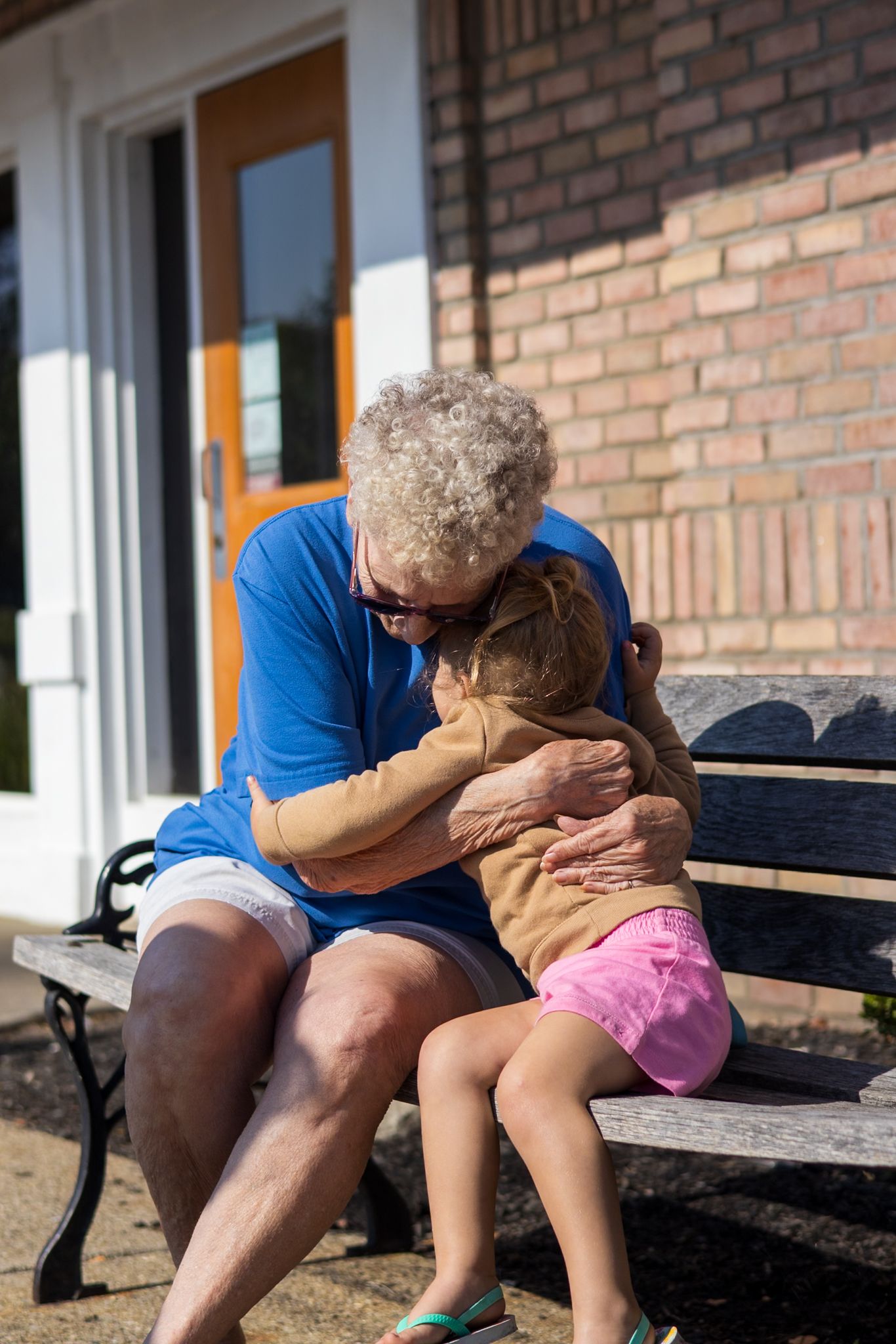 Elderly woman sitting on bench hugging young child outside brick building.