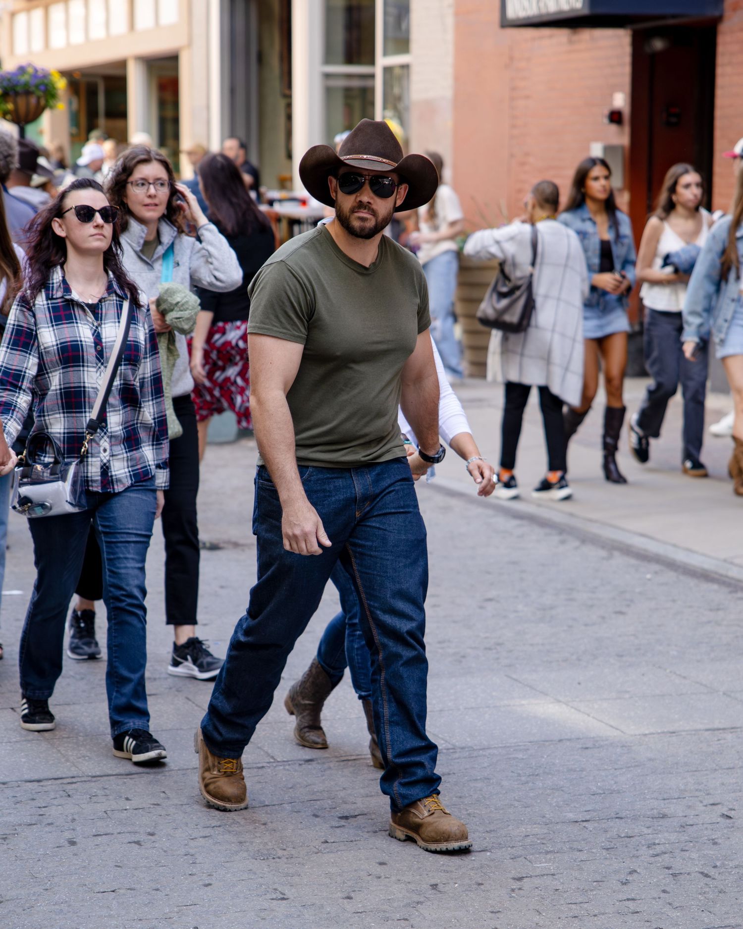 Man in cowboy hat, sunglasses, olive shirt, and jeans walks confidently down a busy urban street.