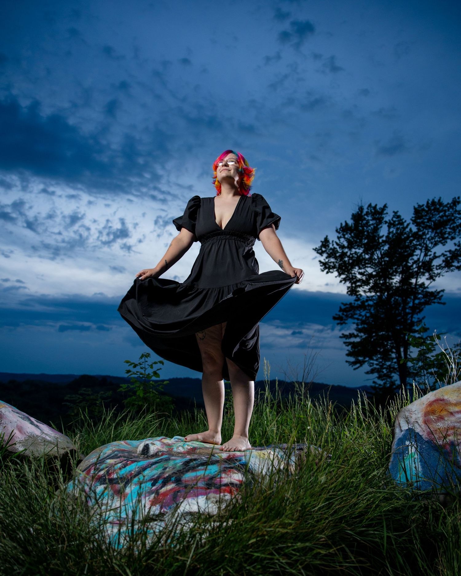 Woman in black dress with red hair stands on painted rocks at dusk under dramatic blue sky.
