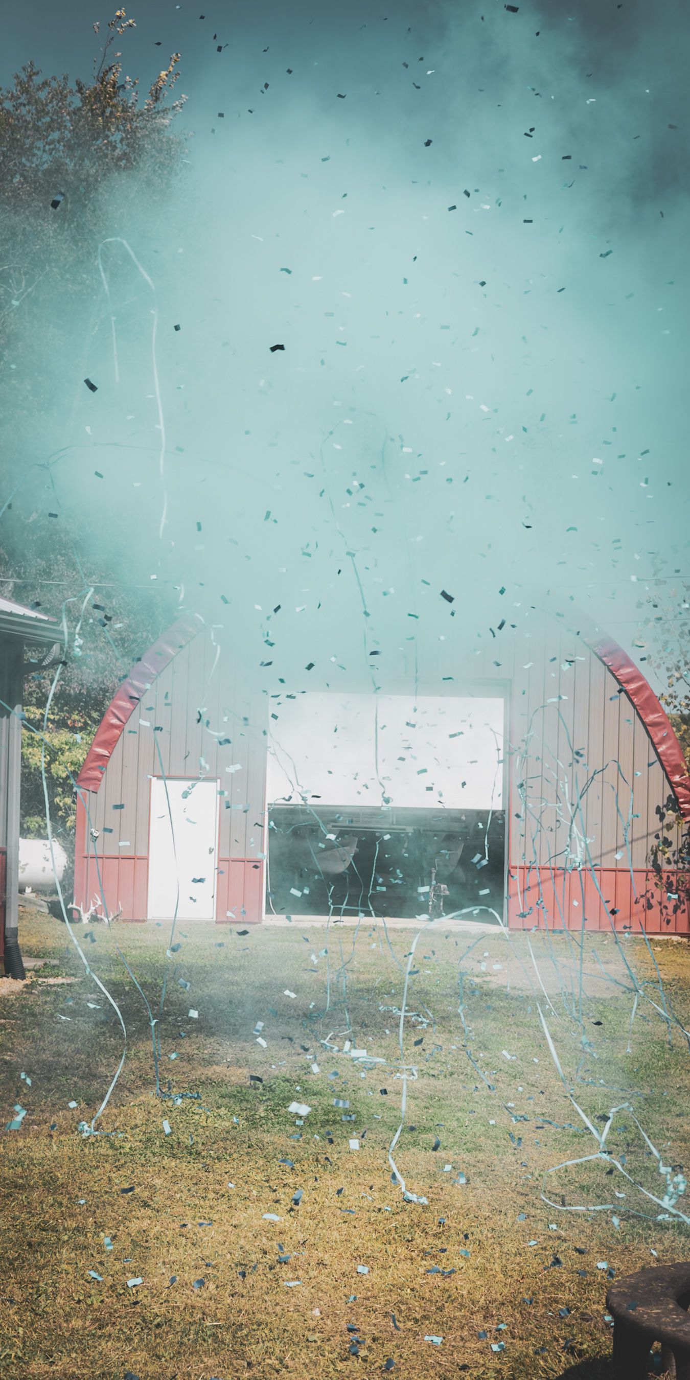 Teal smoke and confetti fill the air at an outdoor gender reveal party revealing a baby boy.