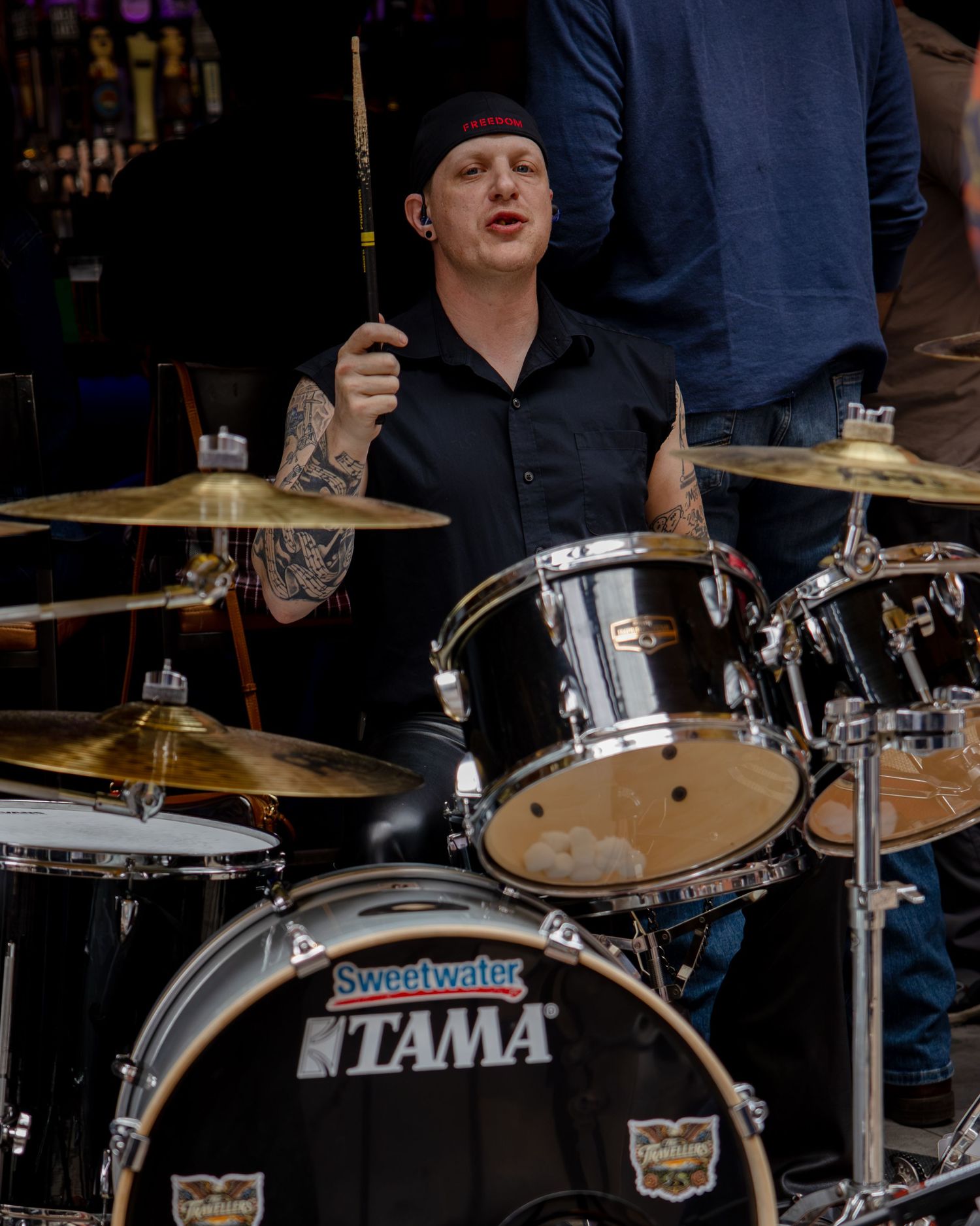 Tattooed drummer playing a black Tama drum kit with Sweetwater branding on an outdoor stage.