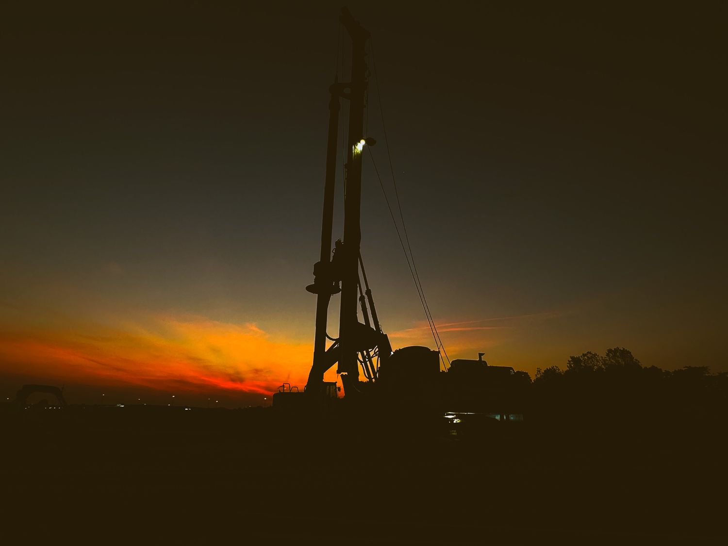 Tall drilling rig silhouetted against a vivid orange and red sunset sky at an outdoor job site.