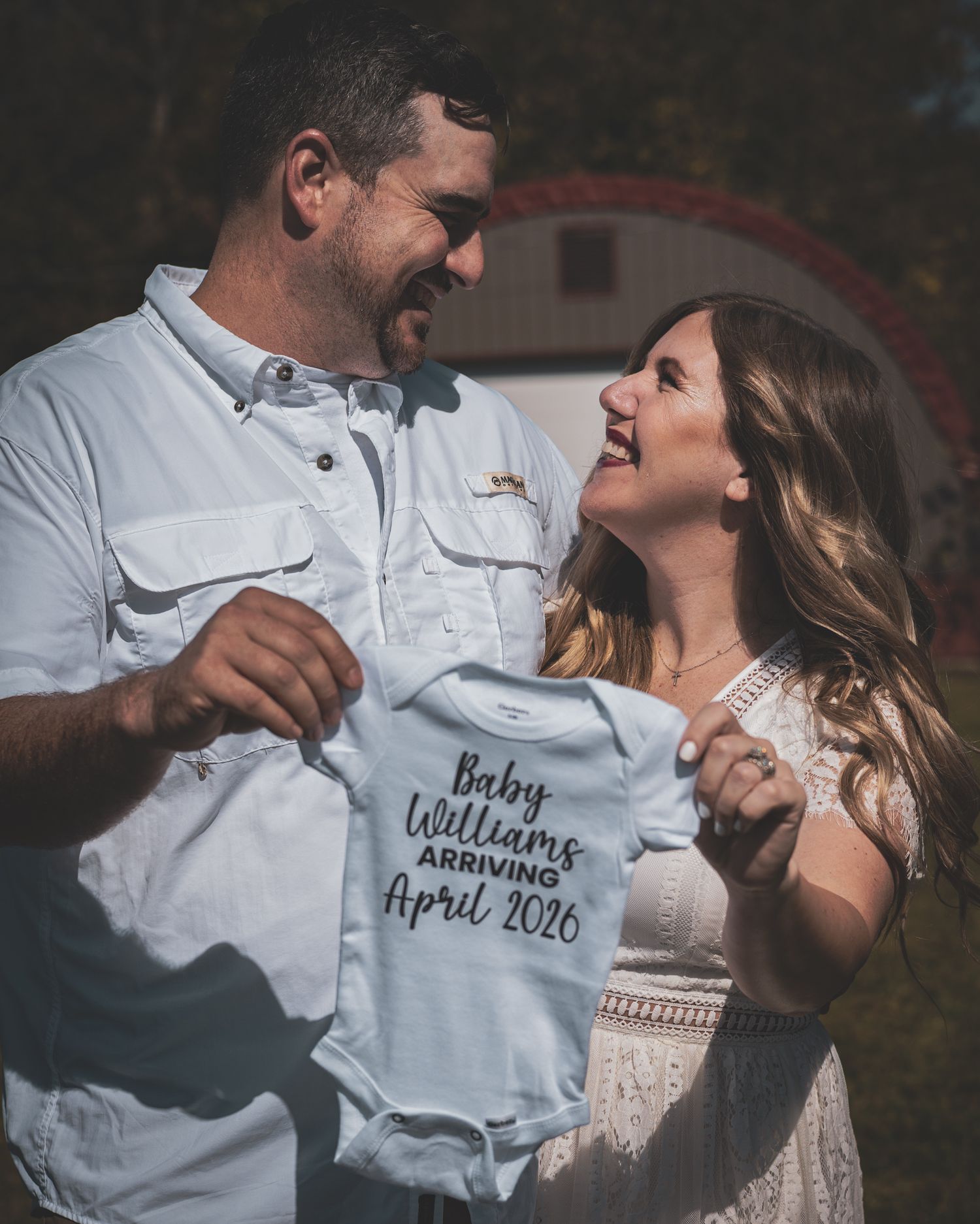 Happy couple holding Baby Williams April 2026 onesie for pregnancy announcement outdoors.