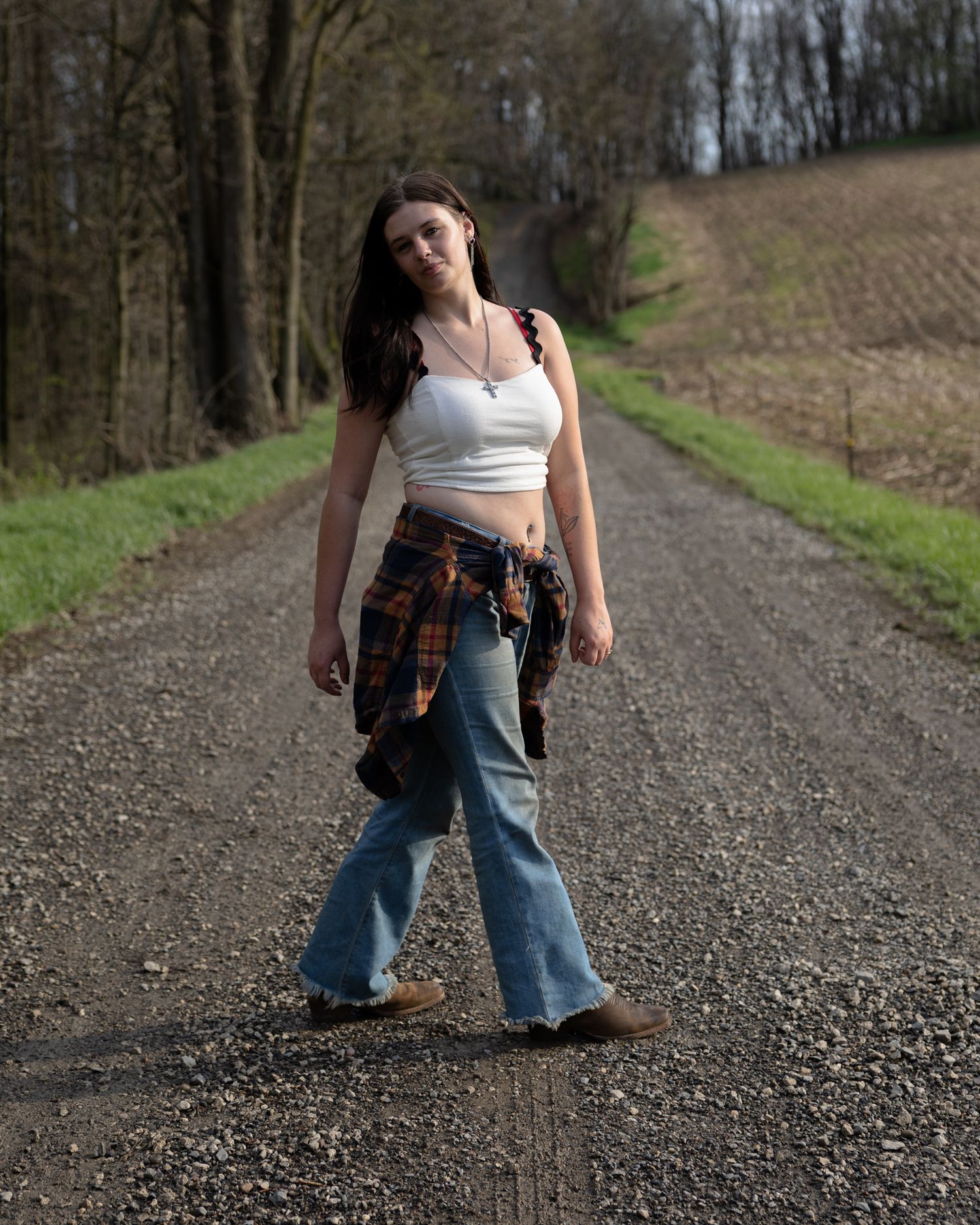 Young woman in white crop top and jeans walking on gravel country road.