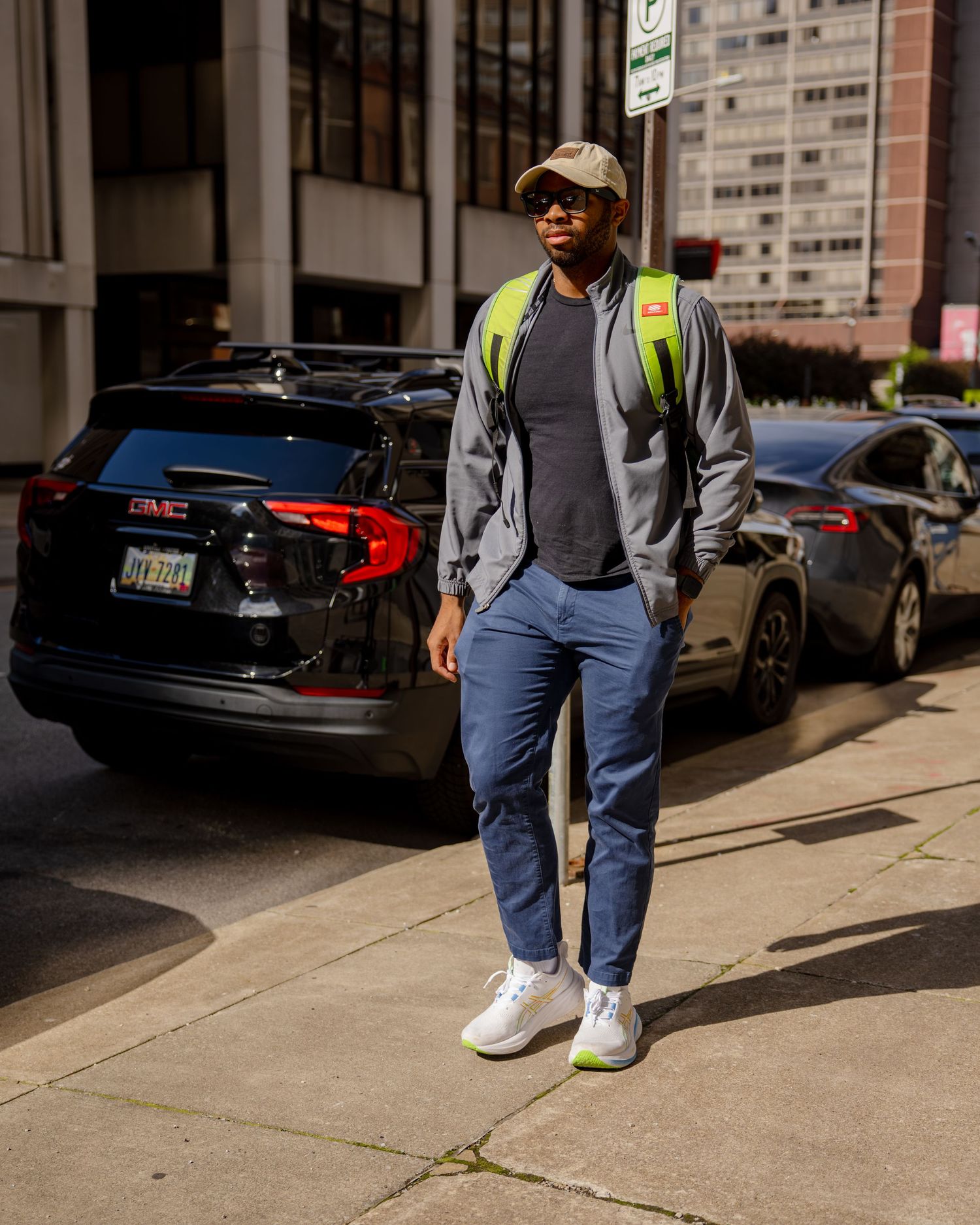 Man in gray jacket, blue pants, cap, and neon green backpack standing on urban sidewalk near parked GMC SUV.