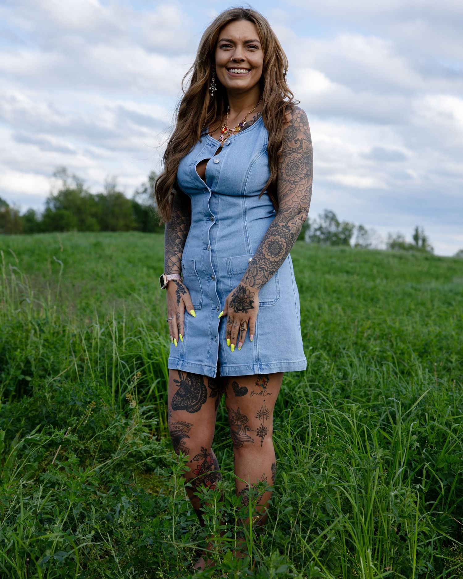 Tattooed woman in short denim dress standing confidently in a lush green field under cloudy sky.