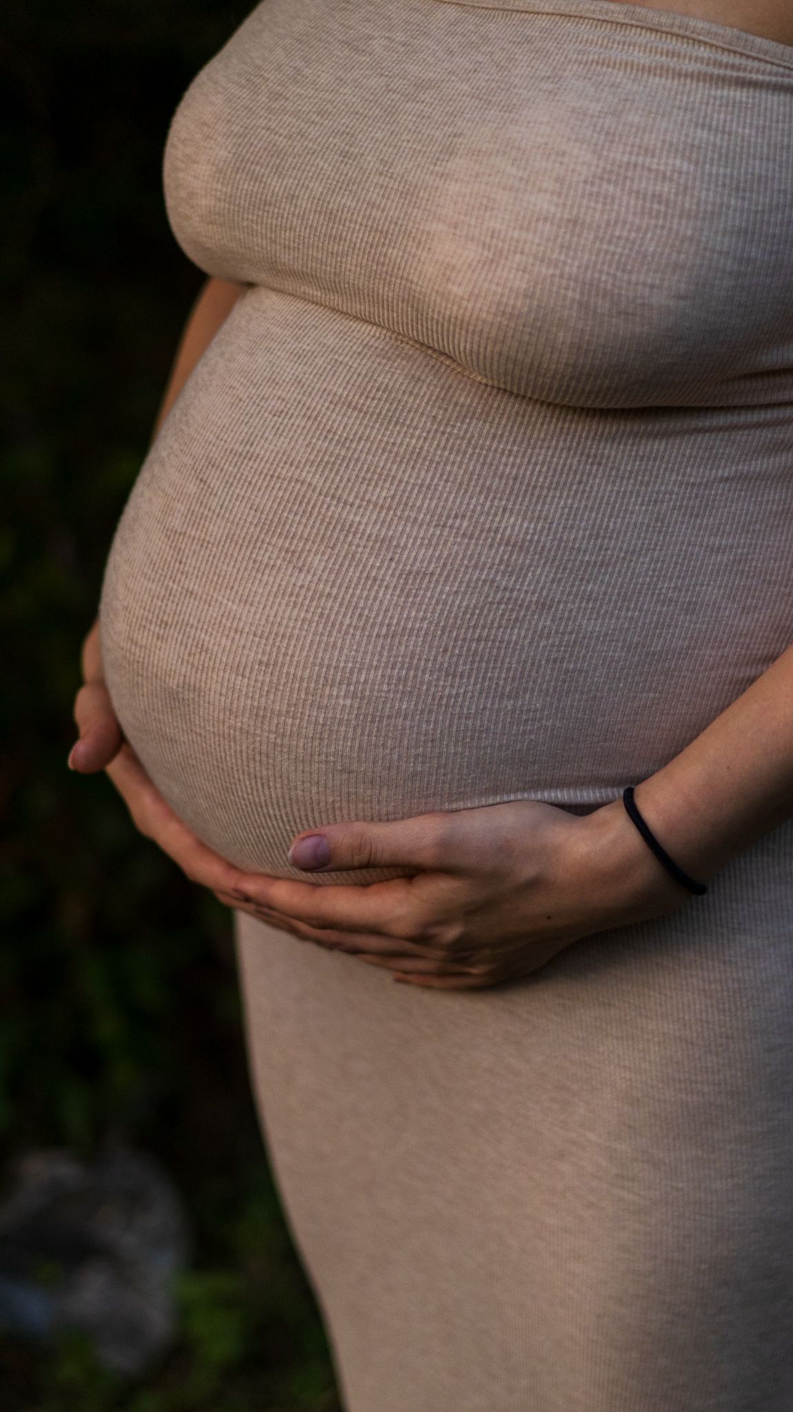 Pregnant woman in beige ribbed dress cradling her baby bump with both hands outdoors.