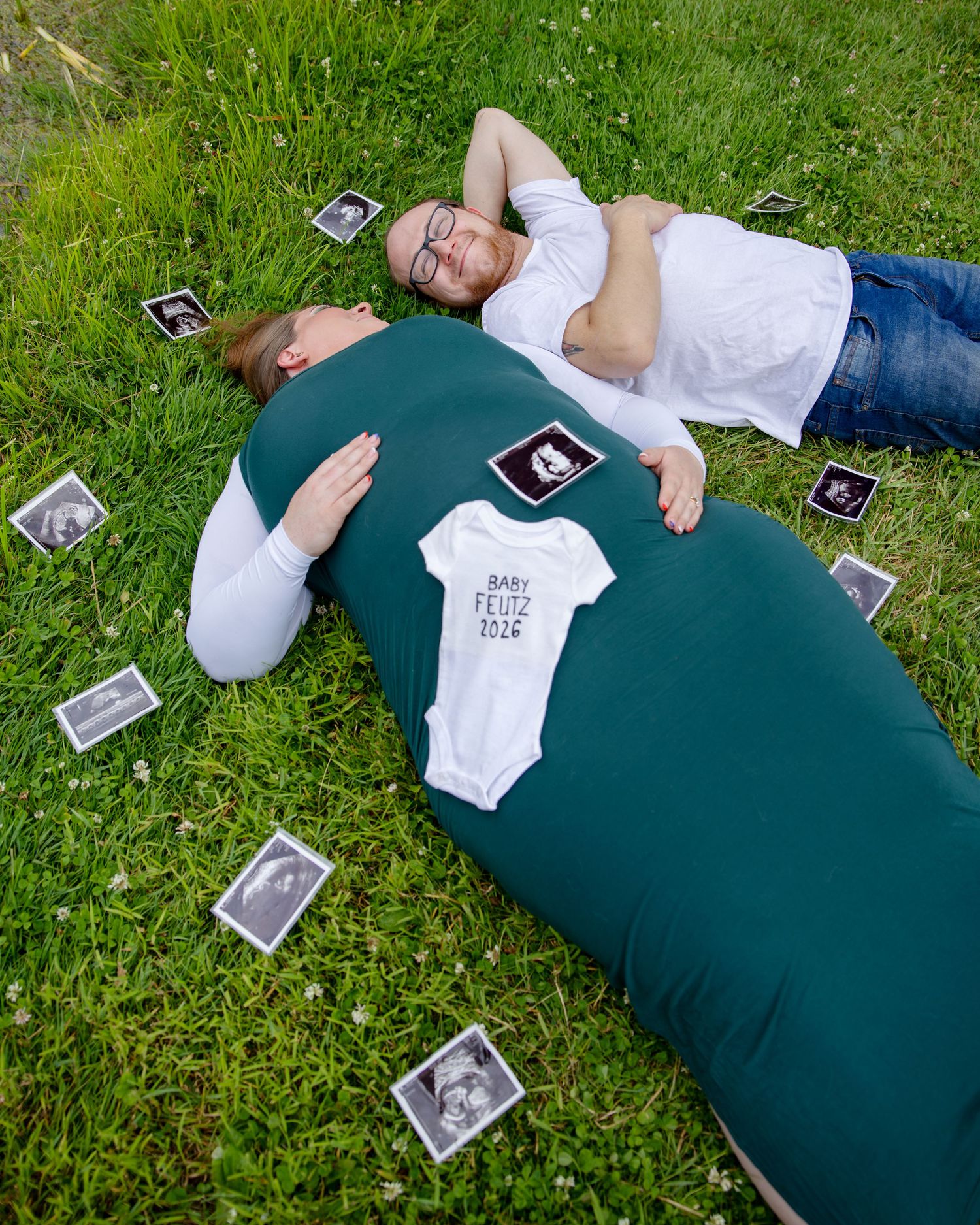 Couple lying on grass with ultrasound photos and baby onesie on pregnant belly for maternity announcement.