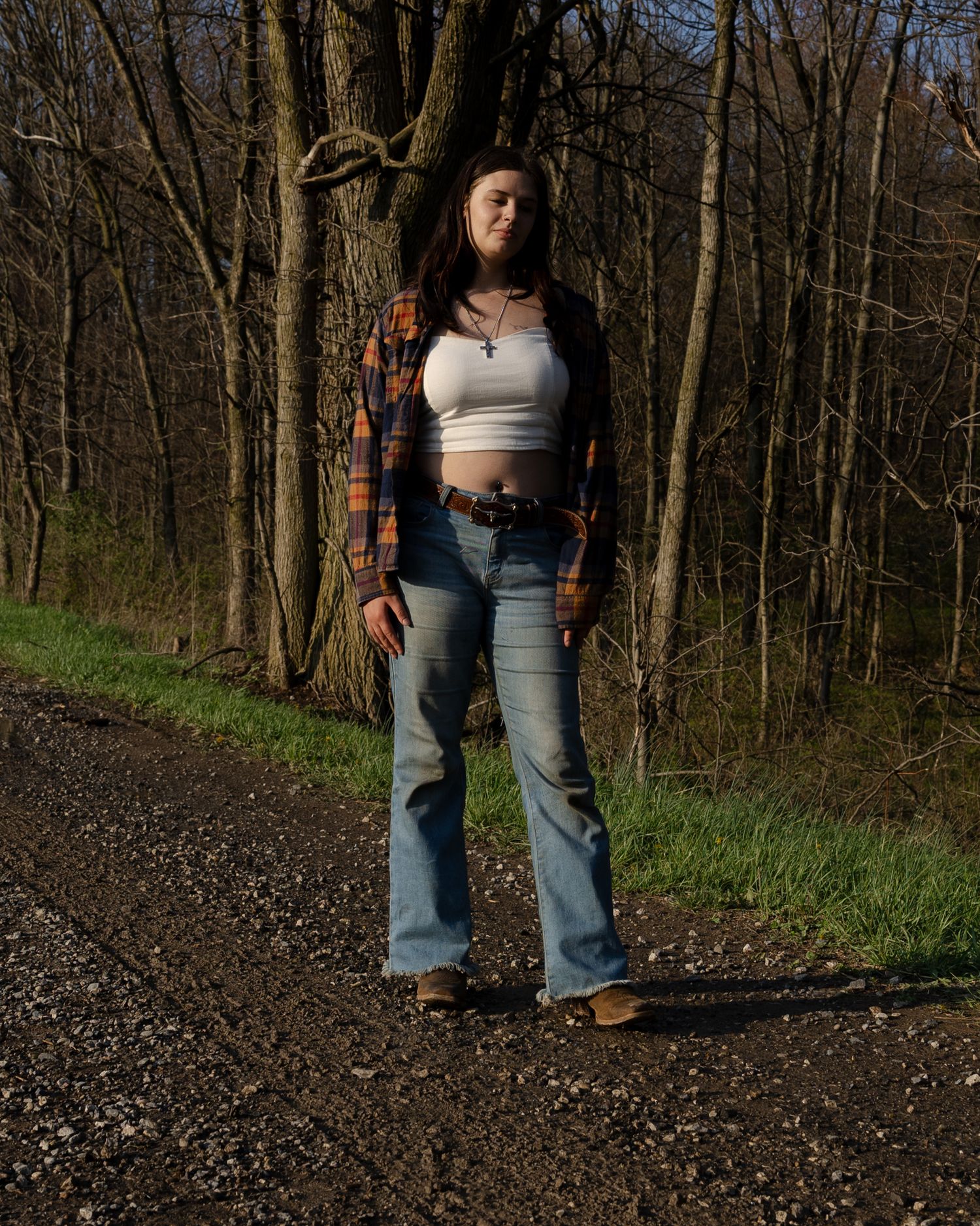 Young woman in white crop top and flared jeans standing on woodland path.