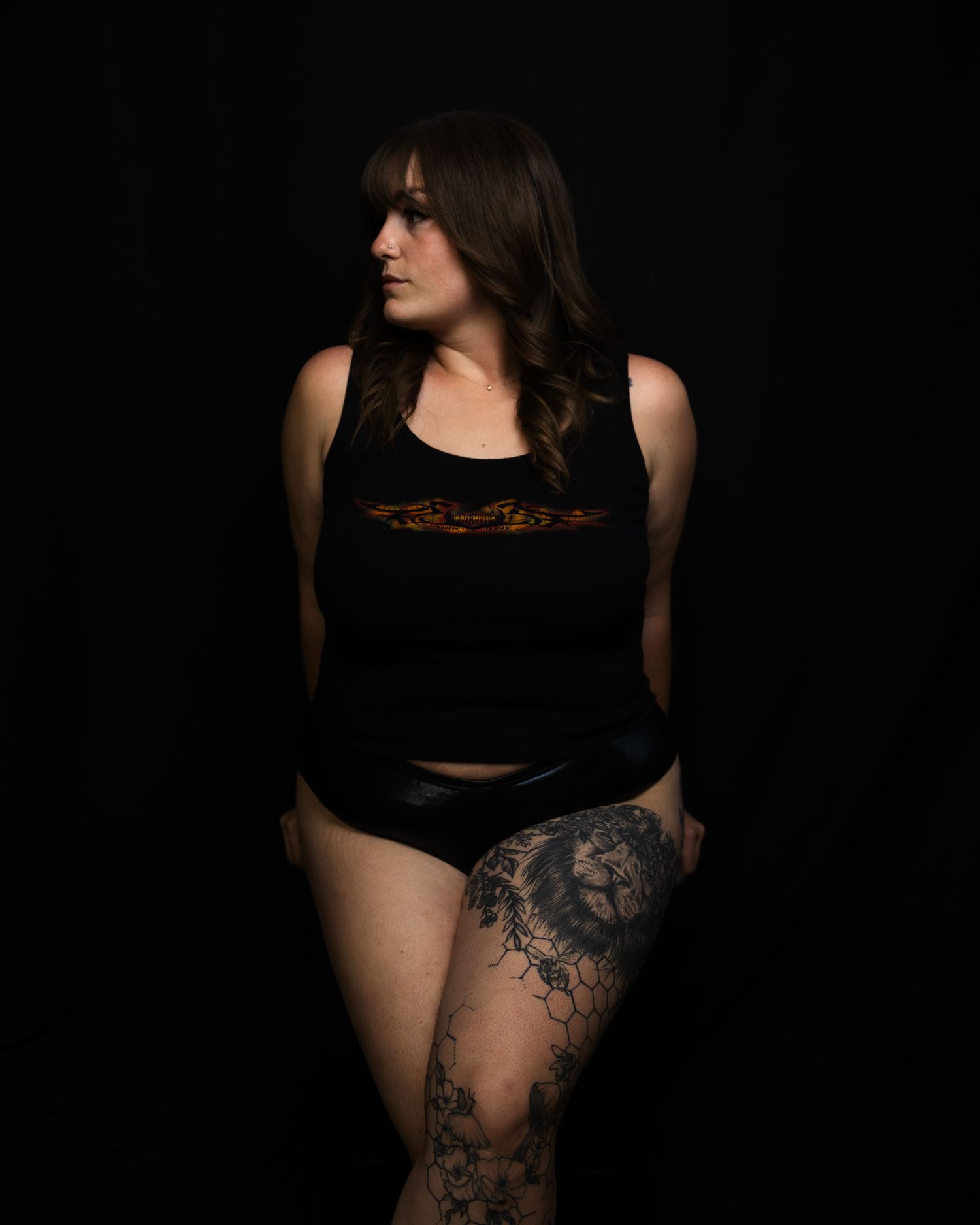 Tattooed brunette woman in black tank top and shorts posing against a dark background.