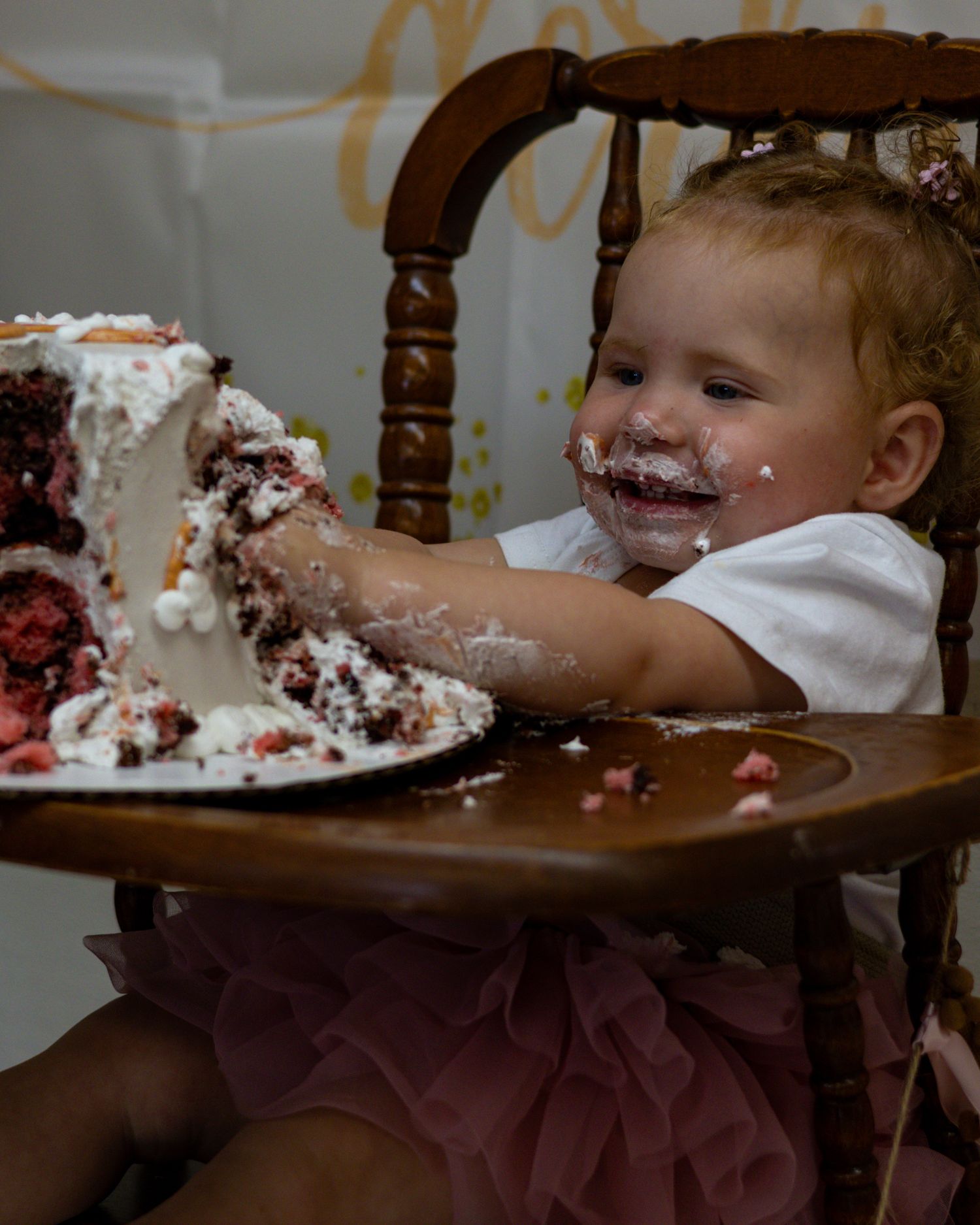 A joyful baby celebrates their first birthday smashing a red velvet cake in a wooden high chair, face covered in frosting.