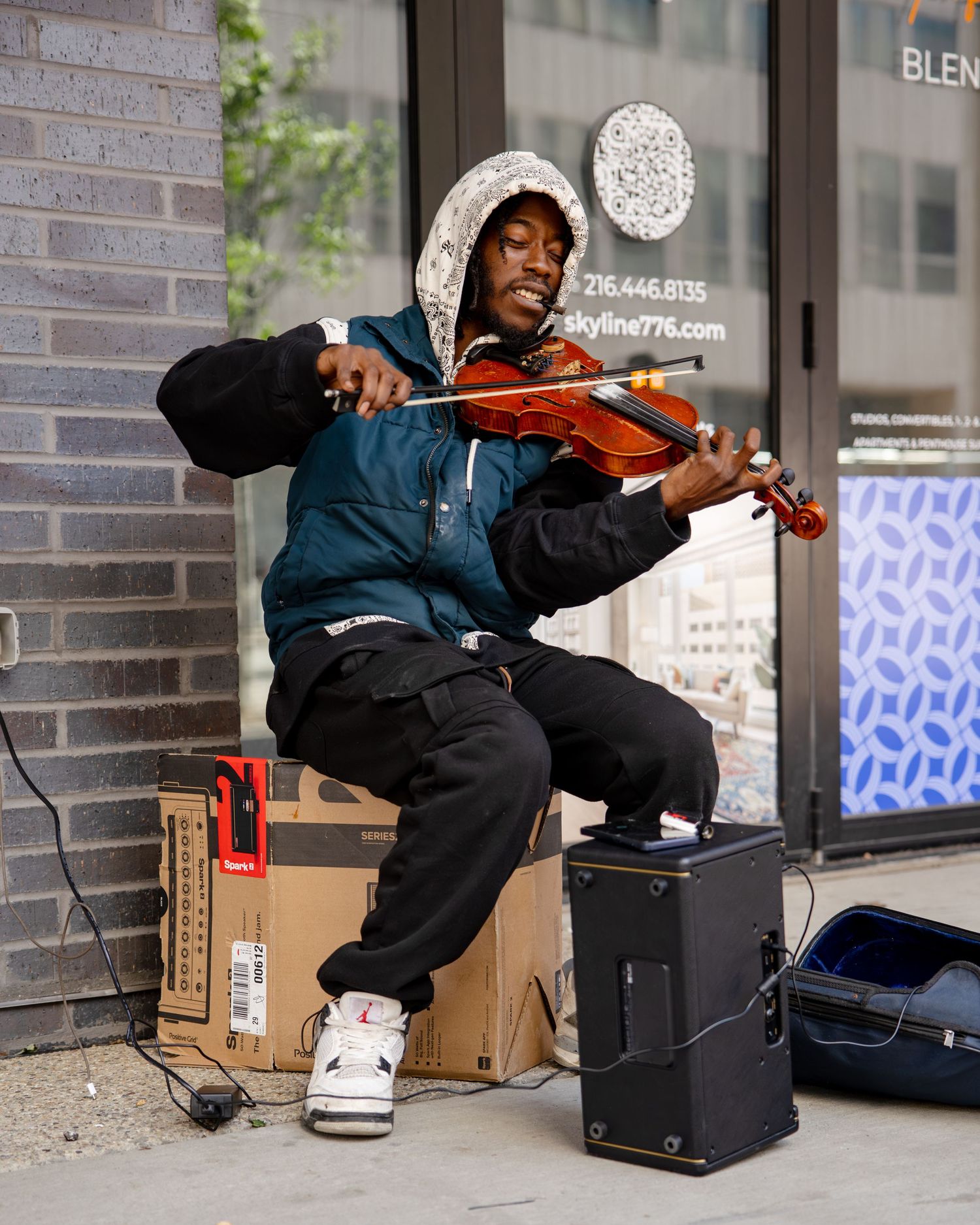 Young musician playing violin while seated on a box outdoors, wearing a teal jacket and white sneakers.