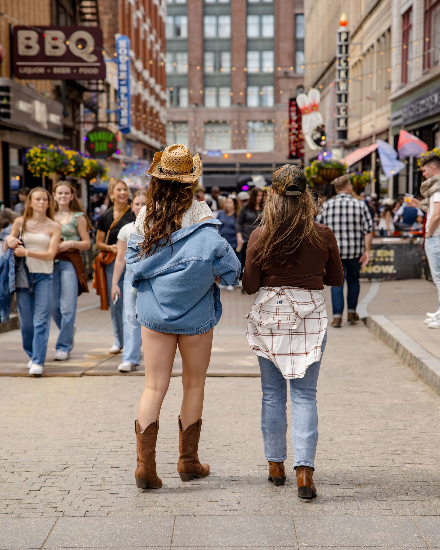 Two women in cowboy boots and hats walk down a busy Nashville street lined with BBQ restaurants and shops.