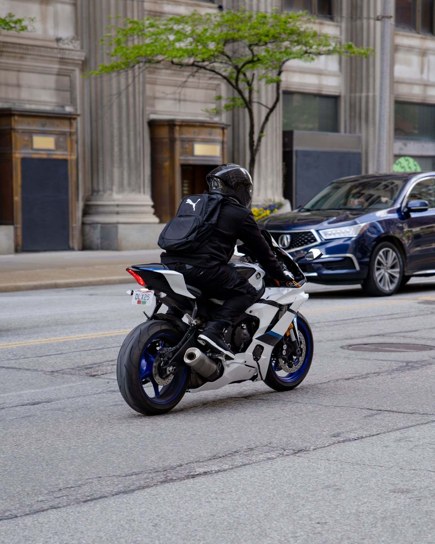 A motorcyclist in black gear and helmet rides a white and blue sport motorcycle through a city street.