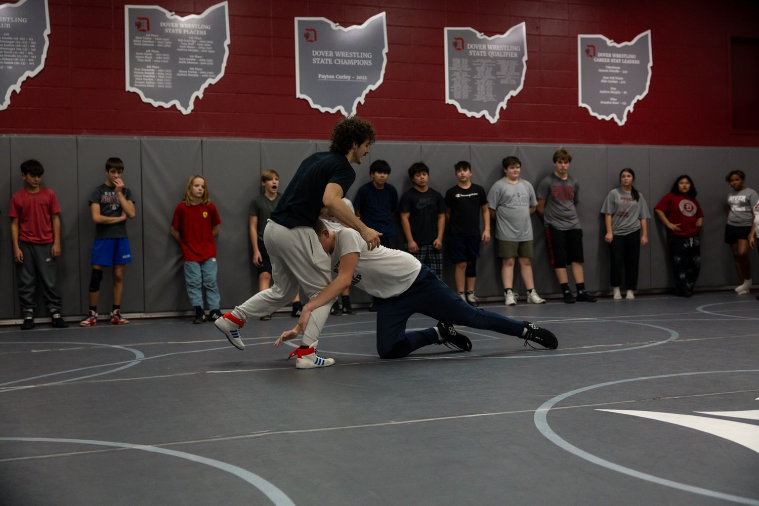 Students watch two wrestlers practice a takedown move on a gym mat in a school wrestling room.