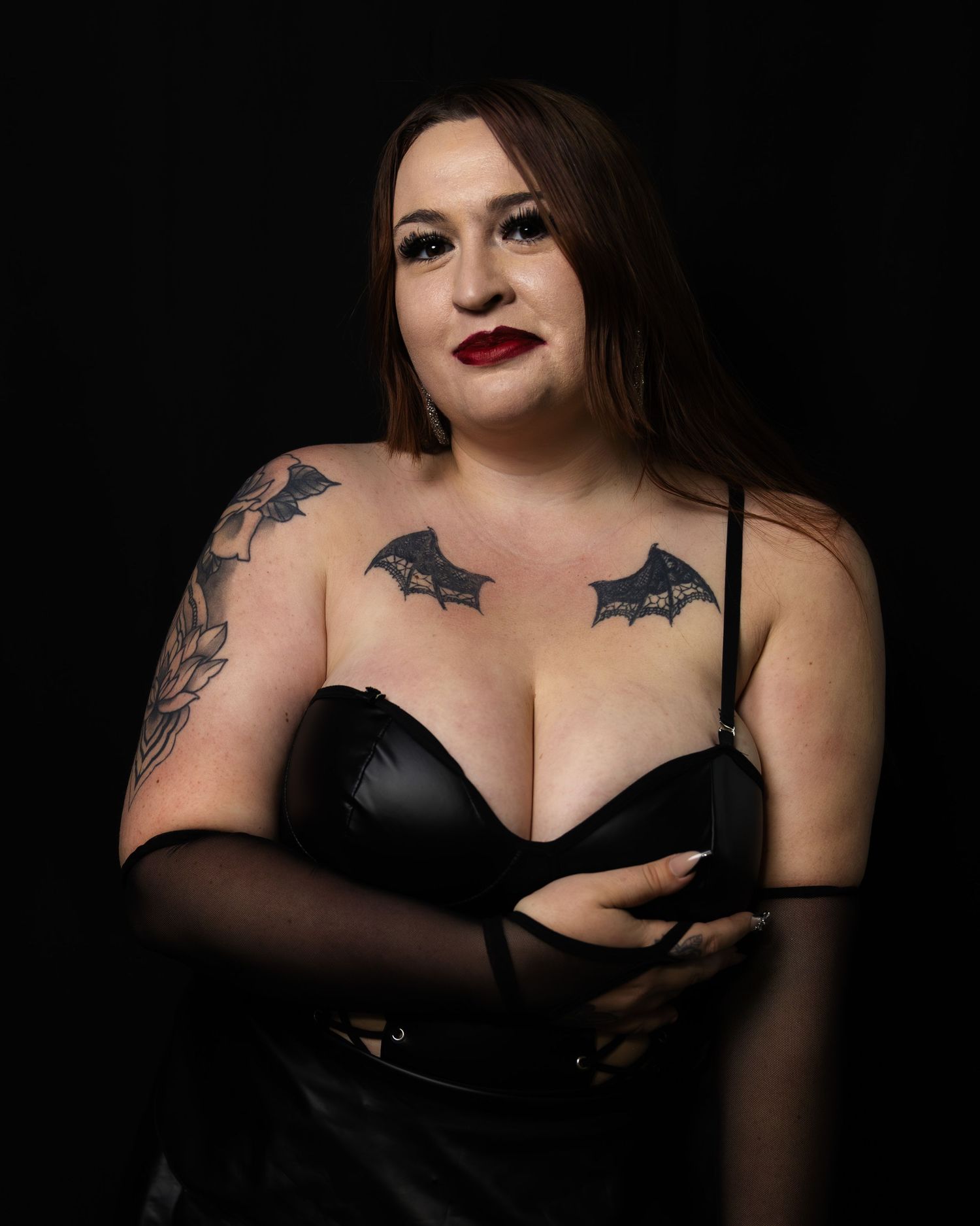 Woman in black leather bustier with bat tattoos posing confidently in studio lighting.
